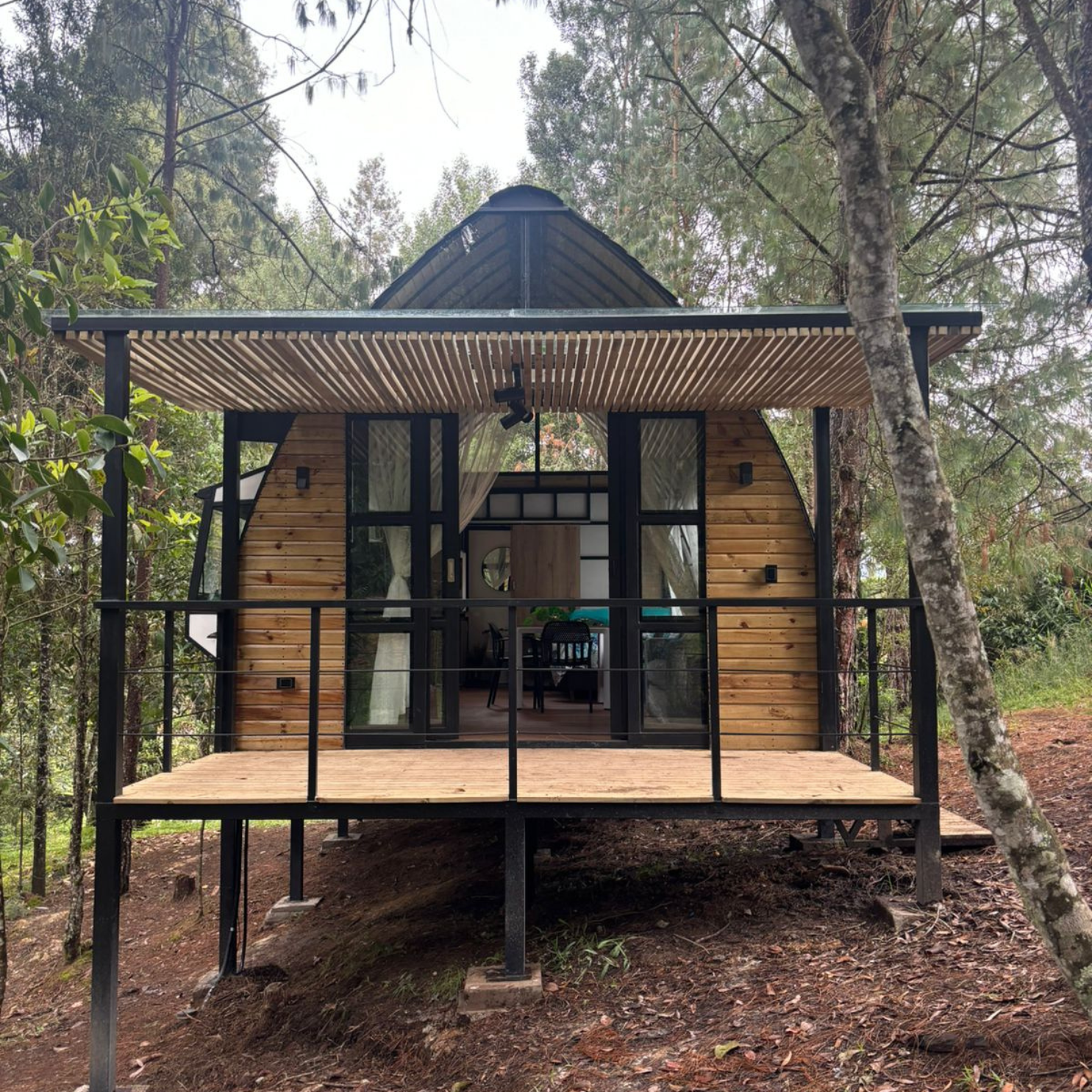 Cabaña moderna de madera con terraza elevada y ventanales en bosque de árboles verdes.