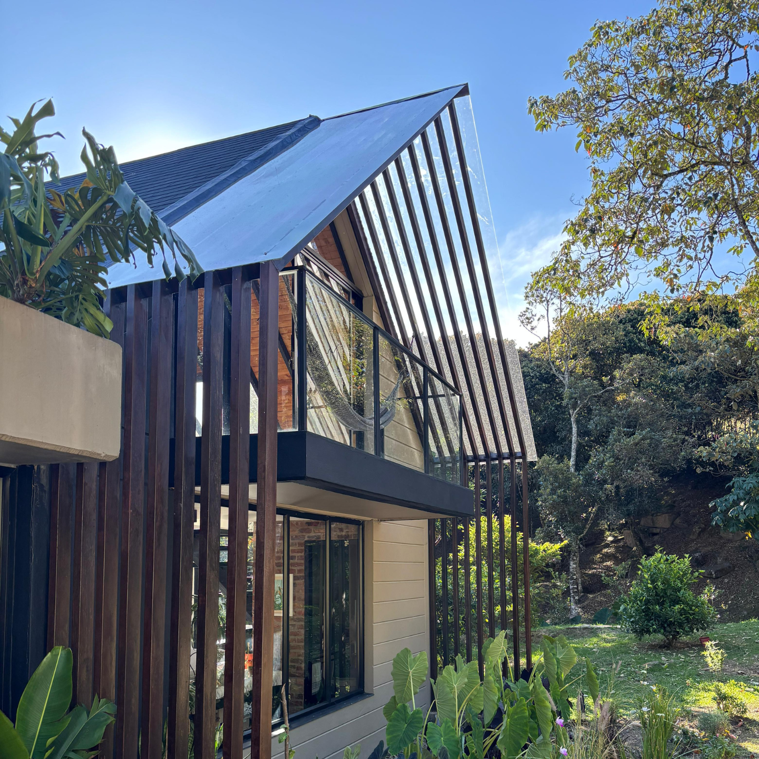 Casa moderna con techo triangular, balcón con hamaca y jardín verde rodeado de árboles.