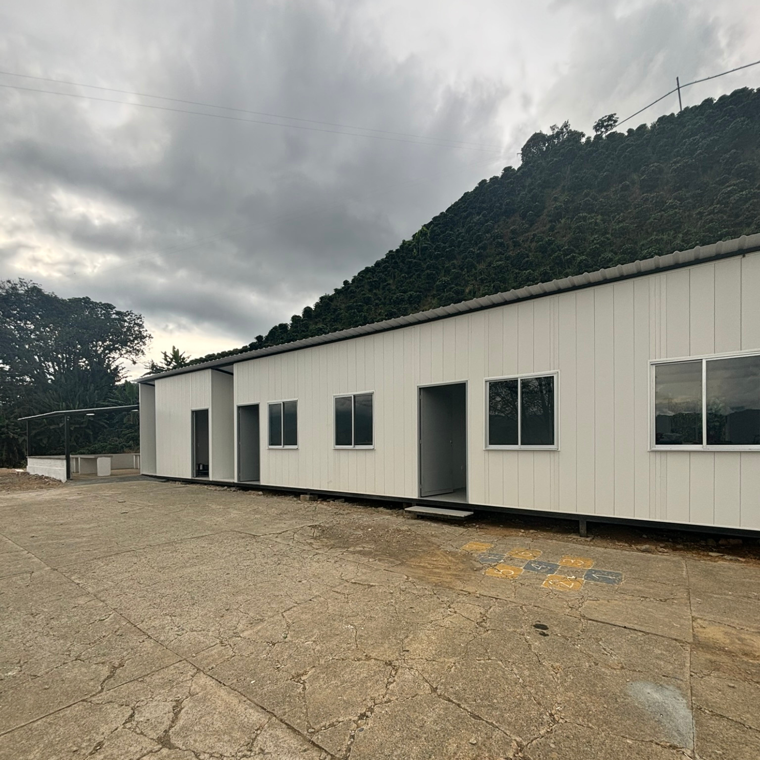 Edificio blanco largo con varias puertas y ventanas, frente a un terreno agrietado y colina verde al fondo.