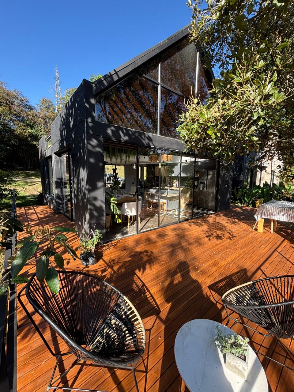 Casa moderna con gran terraza de madera, sillas negras y una mesa blanca con planta bajo un árbol.