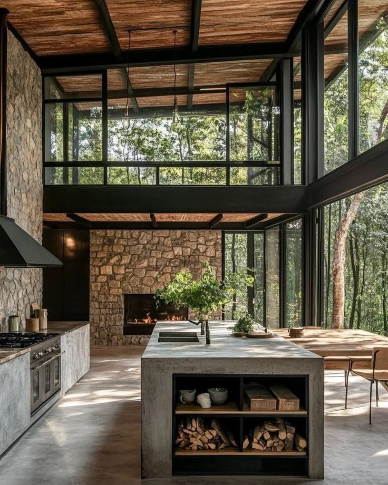 Cocina moderna con isla de concreto, chimenea de piedra y grandes ventanales con vista al bosque.