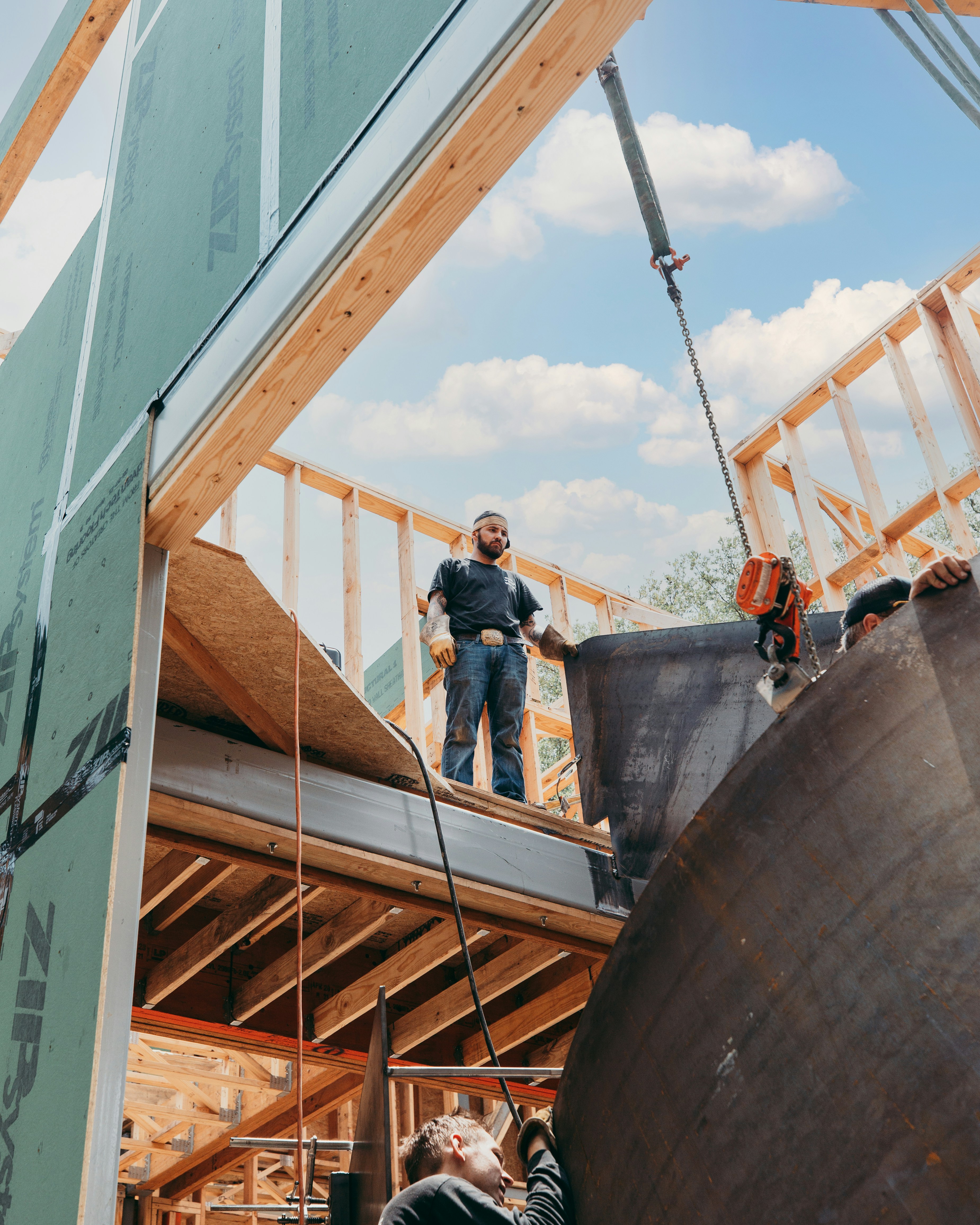 Construction workers installing large metal sheet on a wooden house frame under a blue sky with clouds.