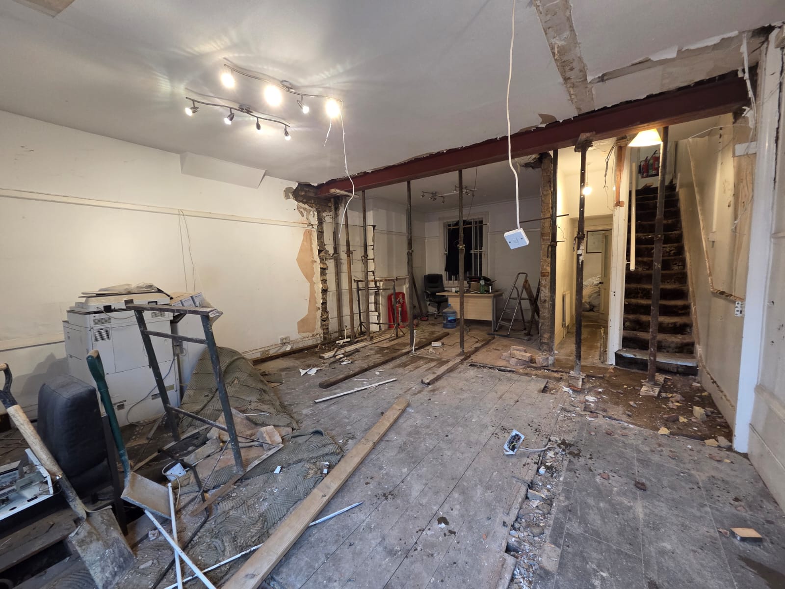 Room under renovation with exposed beams, scattered construction debris, a desk and chair in the back, and a staircase leading up on the right.