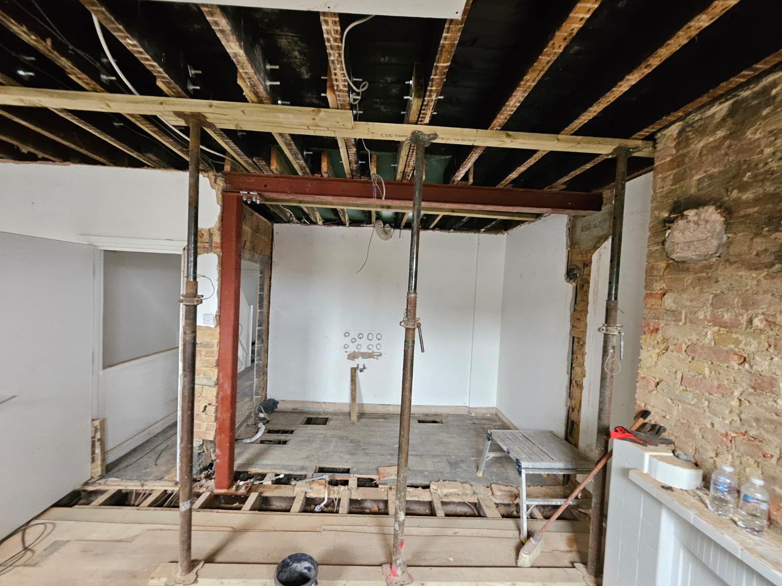 Interior of a room under renovation with exposed ceiling beams, support poles, partially removed floorboards, and construction equipment.