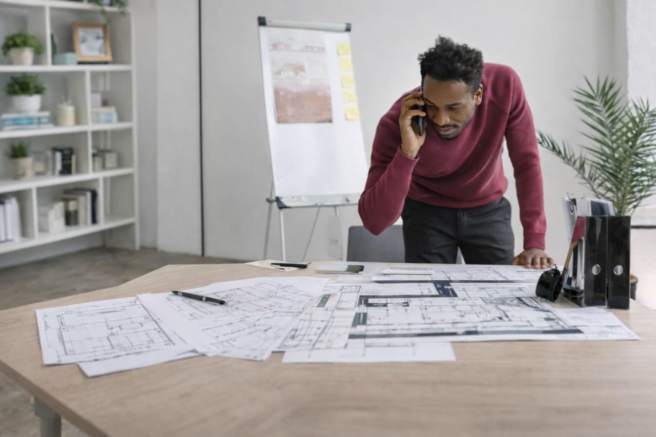 Man in a maroon sweater talking on a phone while reviewing architectural blueprints spread on a table in a modern office.