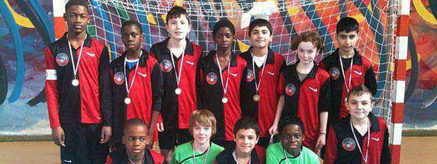 Youth indoor soccer team wearing red and black uniforms posing with medals in front of a goal net.