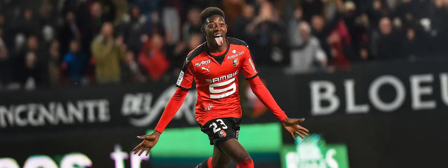 Soccer player in red and black Rennes uniform celebrating with arms outstretched and tongue out during a match.