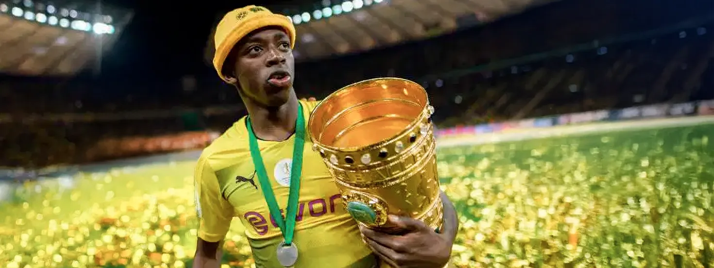 Soccer player wearing a yellow jersey and beanie holding a large gold trophy with confetti on the field.