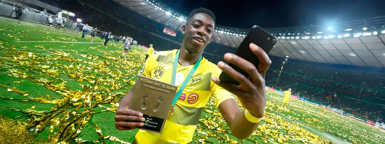 Soccer player in yellow Borussia Dortmund jersey takes a selfie on a confetti-covered field while holding a Man of the Match award plaque.