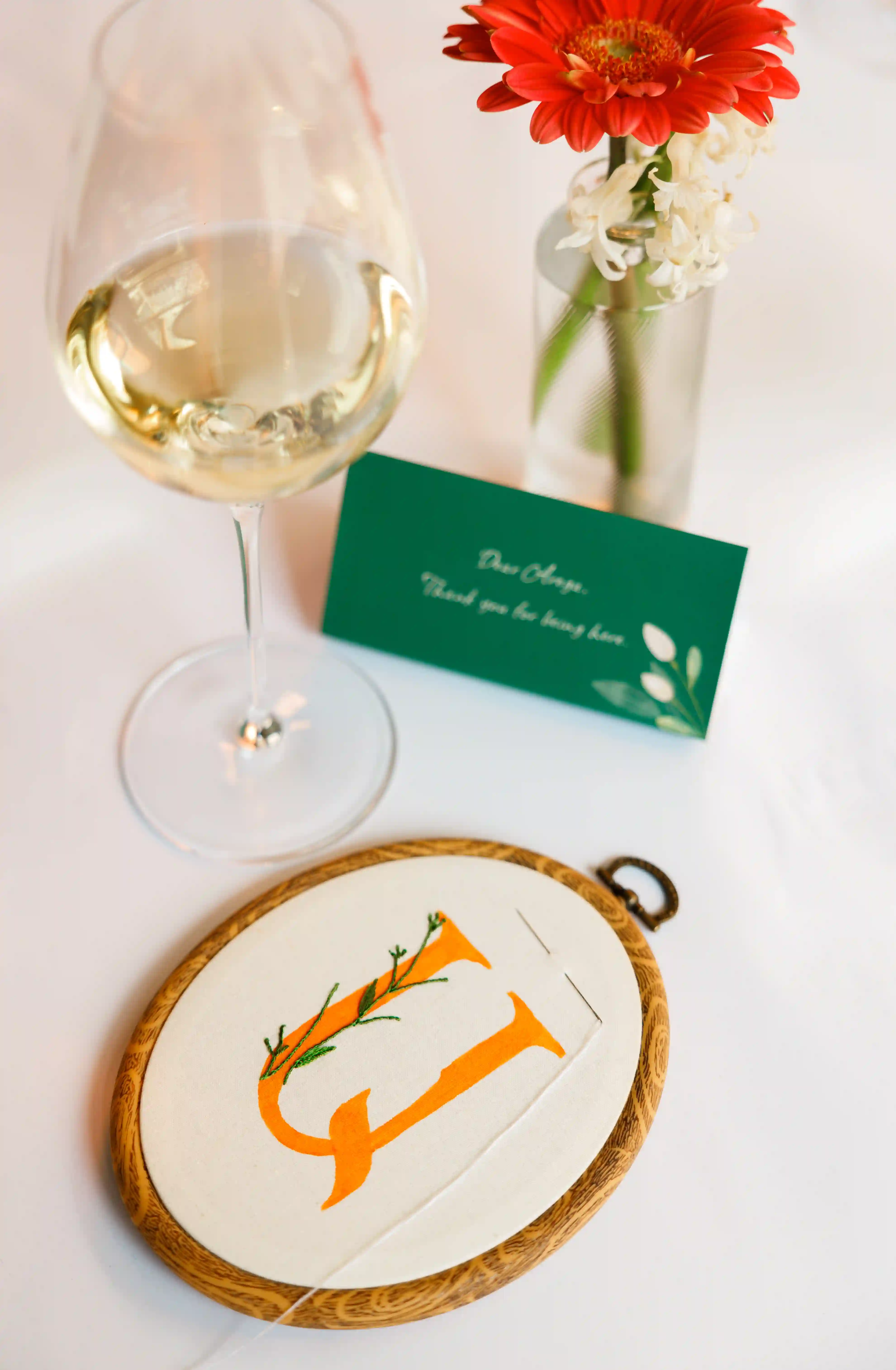Personalized embroidery hoop monogram keepsake beside a white wine glass, green thank you card, and red gerbera bud vase at a luxury event place setting by Perla Signature Events in Yerevan, Armenia