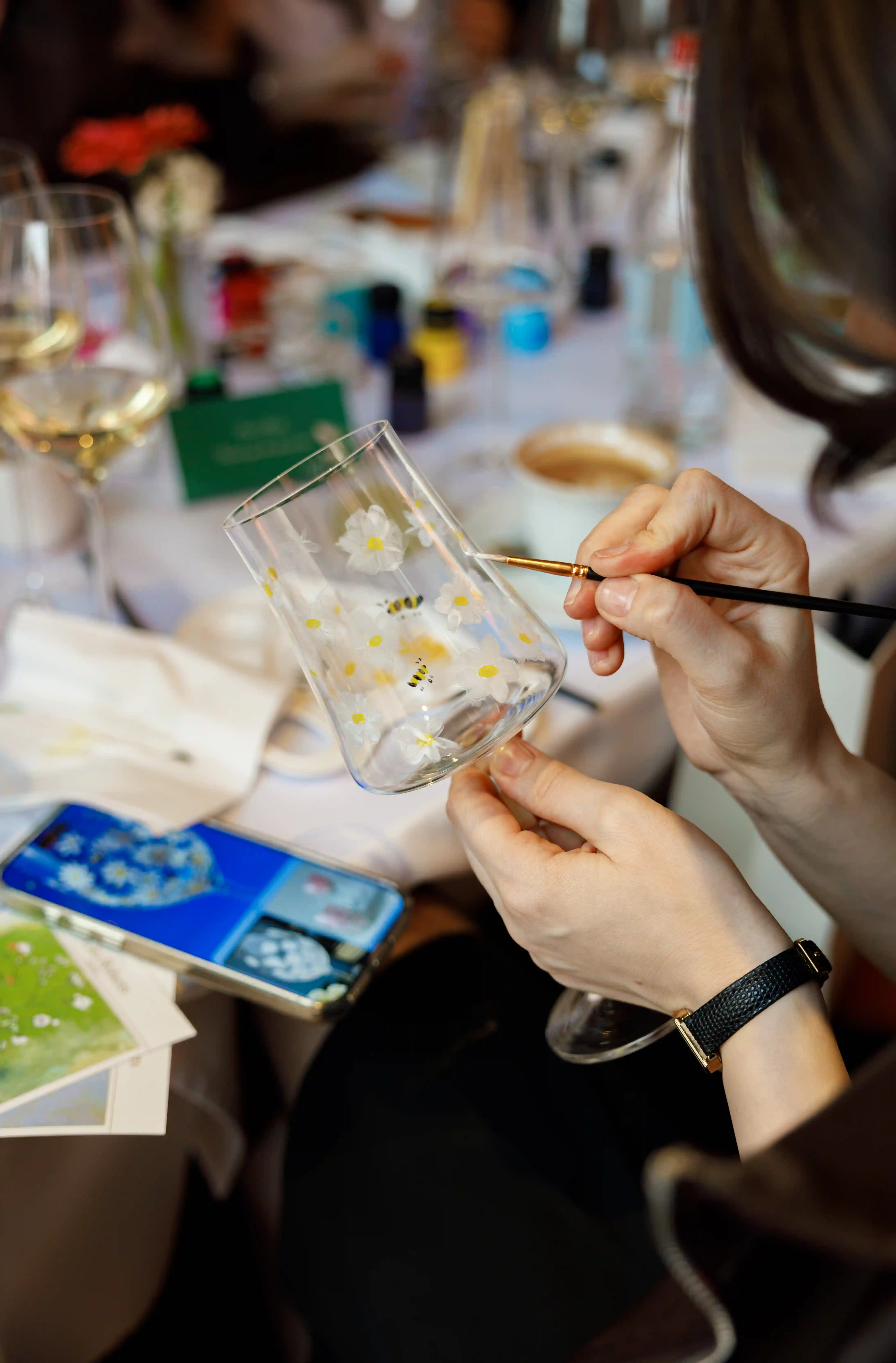 Guest hand-painting white daisy and bee motifs onto a crystal glass with a fine brush at a luxury glass painting workshop event by Perla Signature Events in Yerevan, Armenia