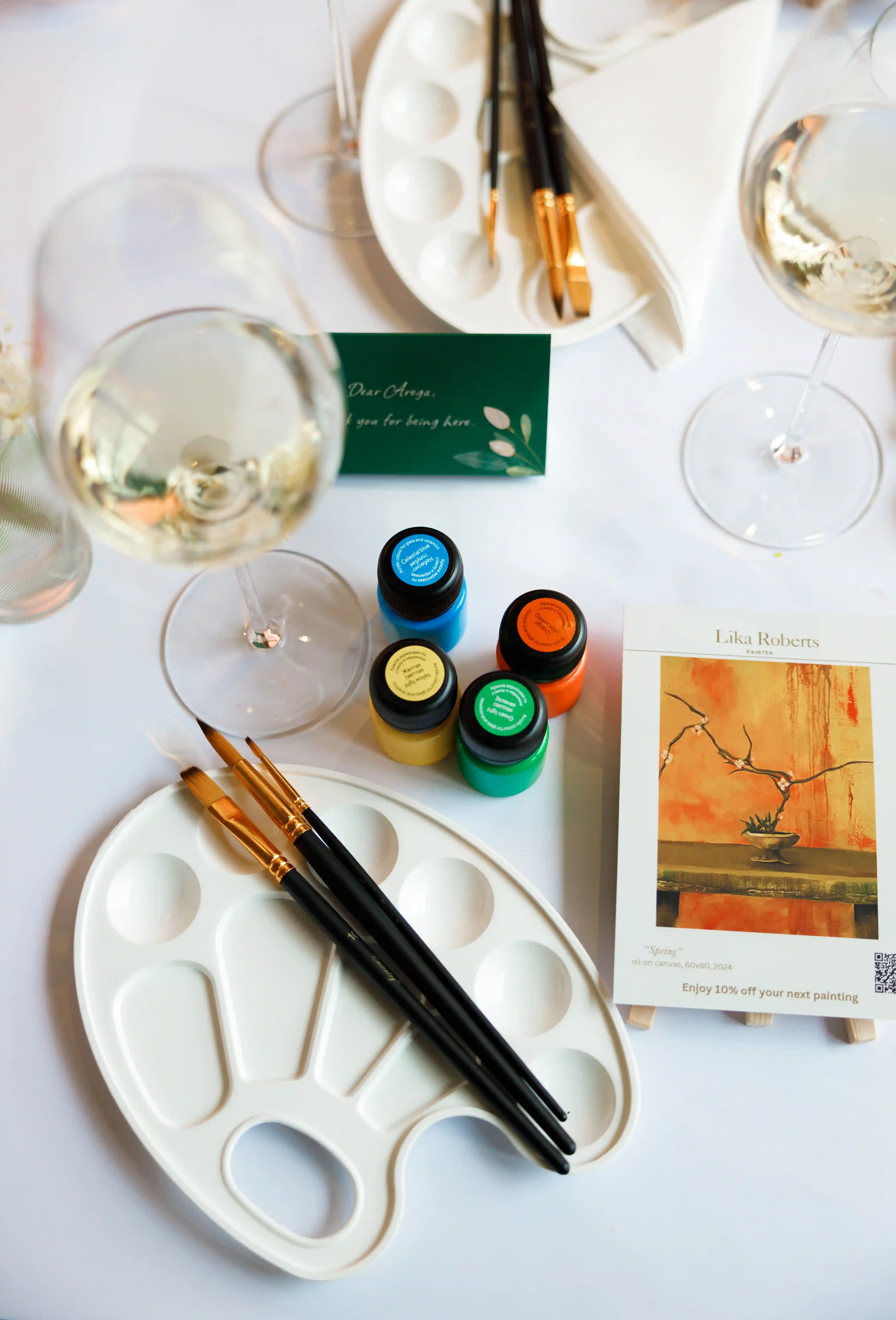 Luxury paint and sip event table setup with ceramic paint palettes, gold-handled brushes, colourful acrylic paint pots, crystal wine glasses and a personalized green guest card by Perla Signature Events in Yerevan, Armenia