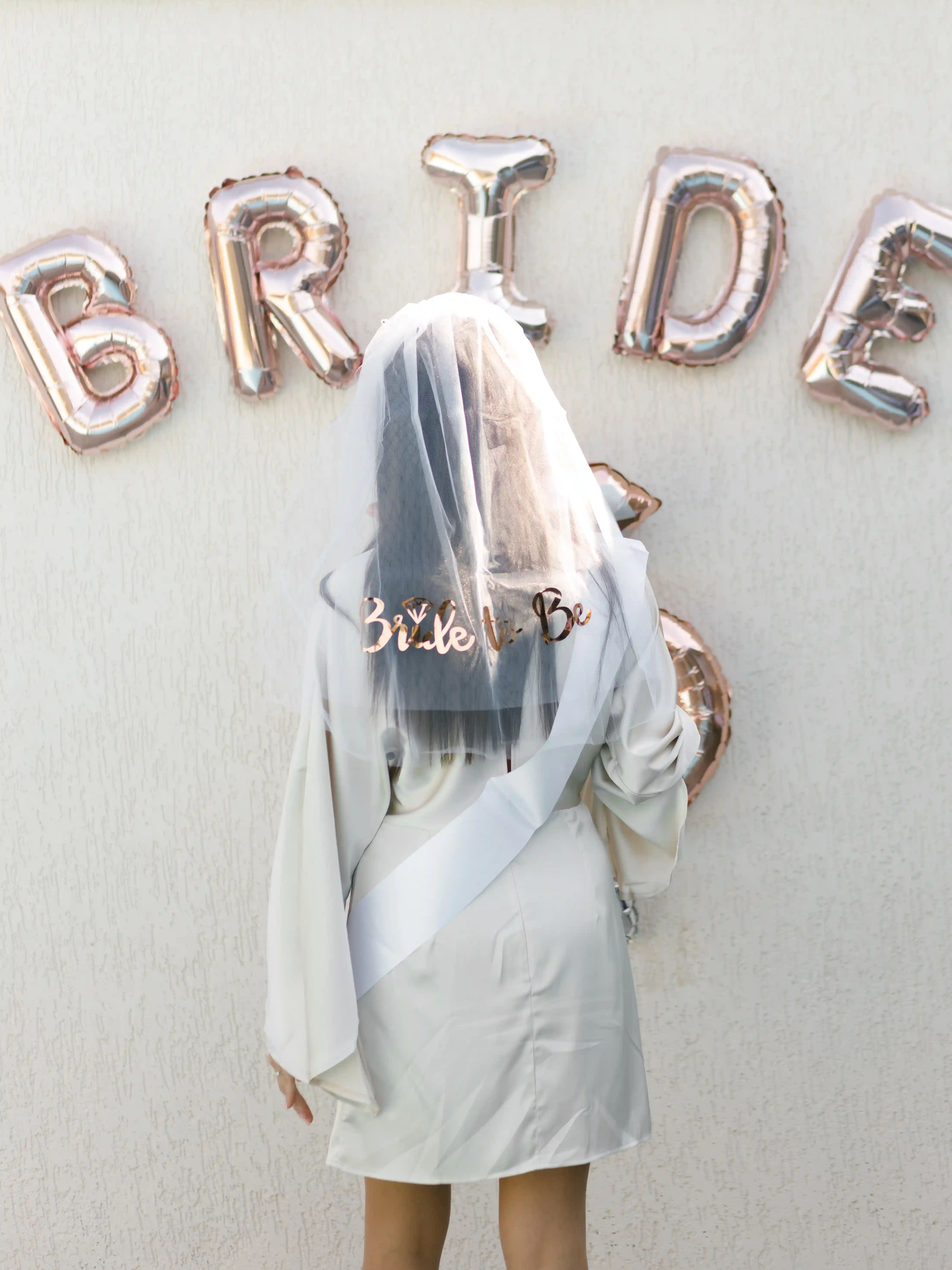 Bride-to-be in a silk robe, `Bride to Be` veil and white sash posing in front of rose gold BRIDE foil balloon letters at a luxury bachelorette party planned by Perla Signature Events in Yerevan, Armenia