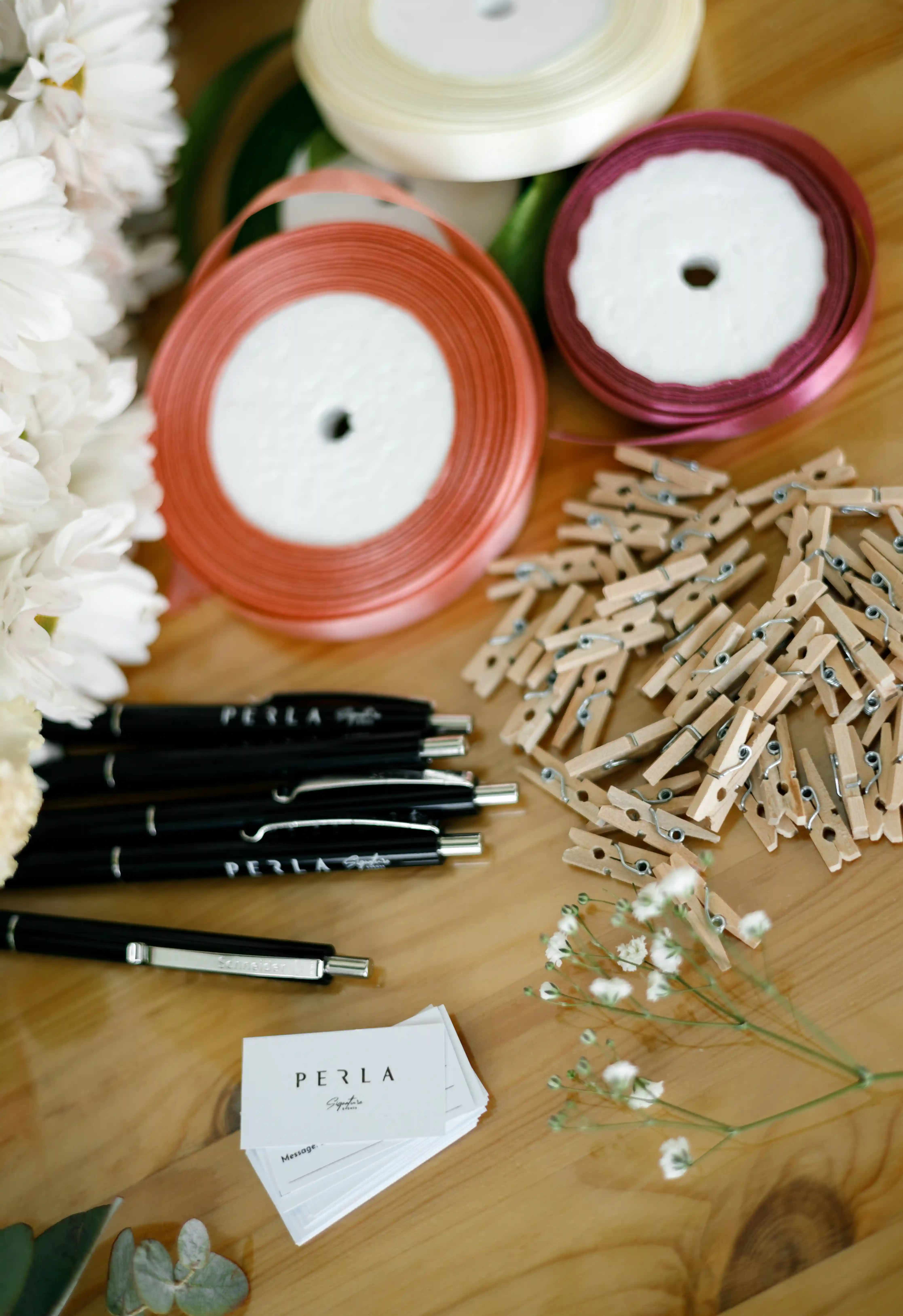 Perla Signature Events branded pens, message cards, mini wooden pegs, satin ribbon rolls and white chrysanthemums laid out as a luxury event preparation flat lay in Yerevan, Armenia