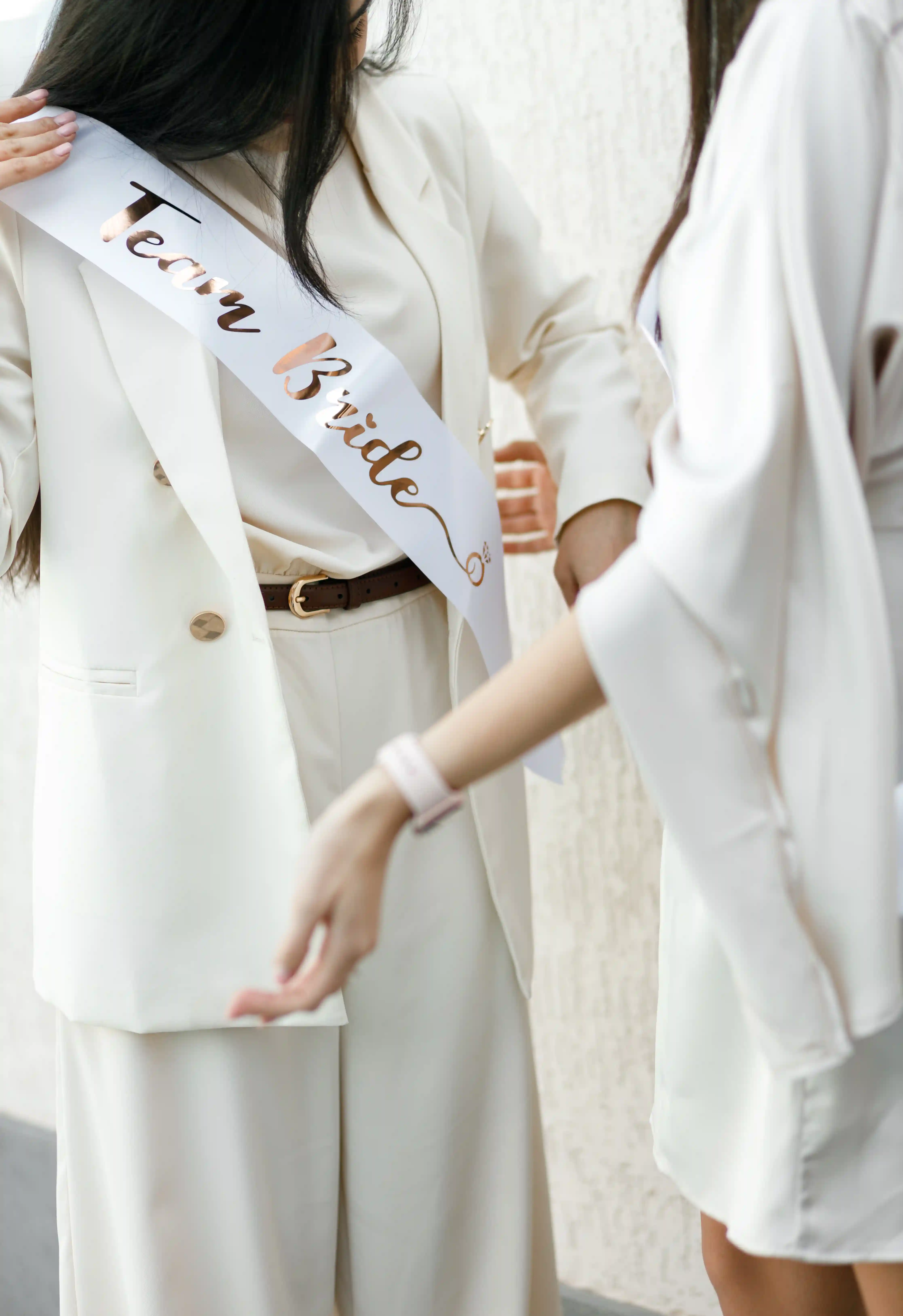 Bridesmaid placing a rose gold Team Bride sash on a guest in a cream blazer and wide-leg trousers at a luxury bridal shower planned by Perla Signature Events in Yerevan, Armenia