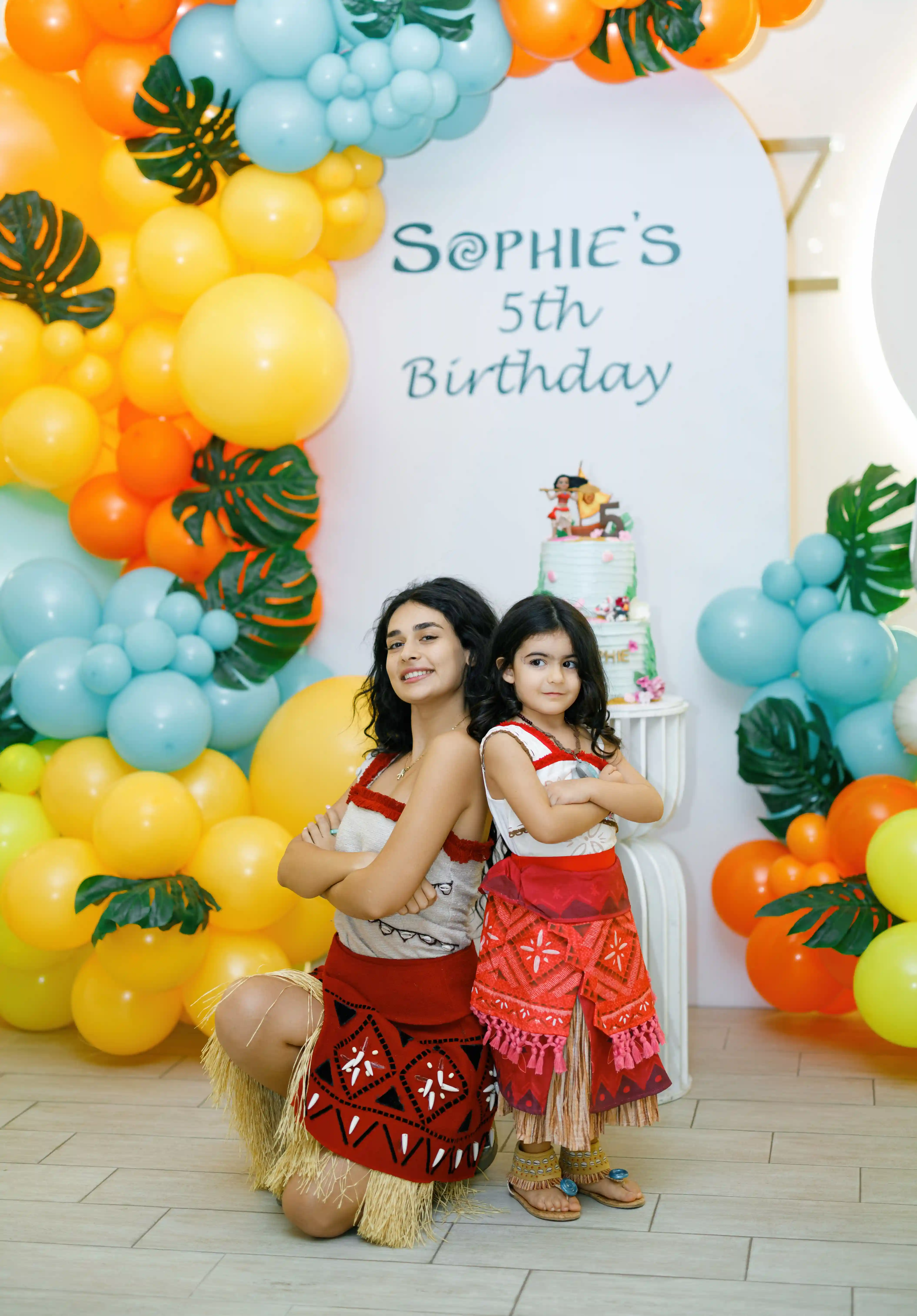 Birthday girl and entertainer in matching tropical island princess costumes posing back-to-back in front of a vibrant orange, yellow, and teal balloon arch at a luxury themed fifth birthday party by Perla Signature Events in Yerevan, Armenia