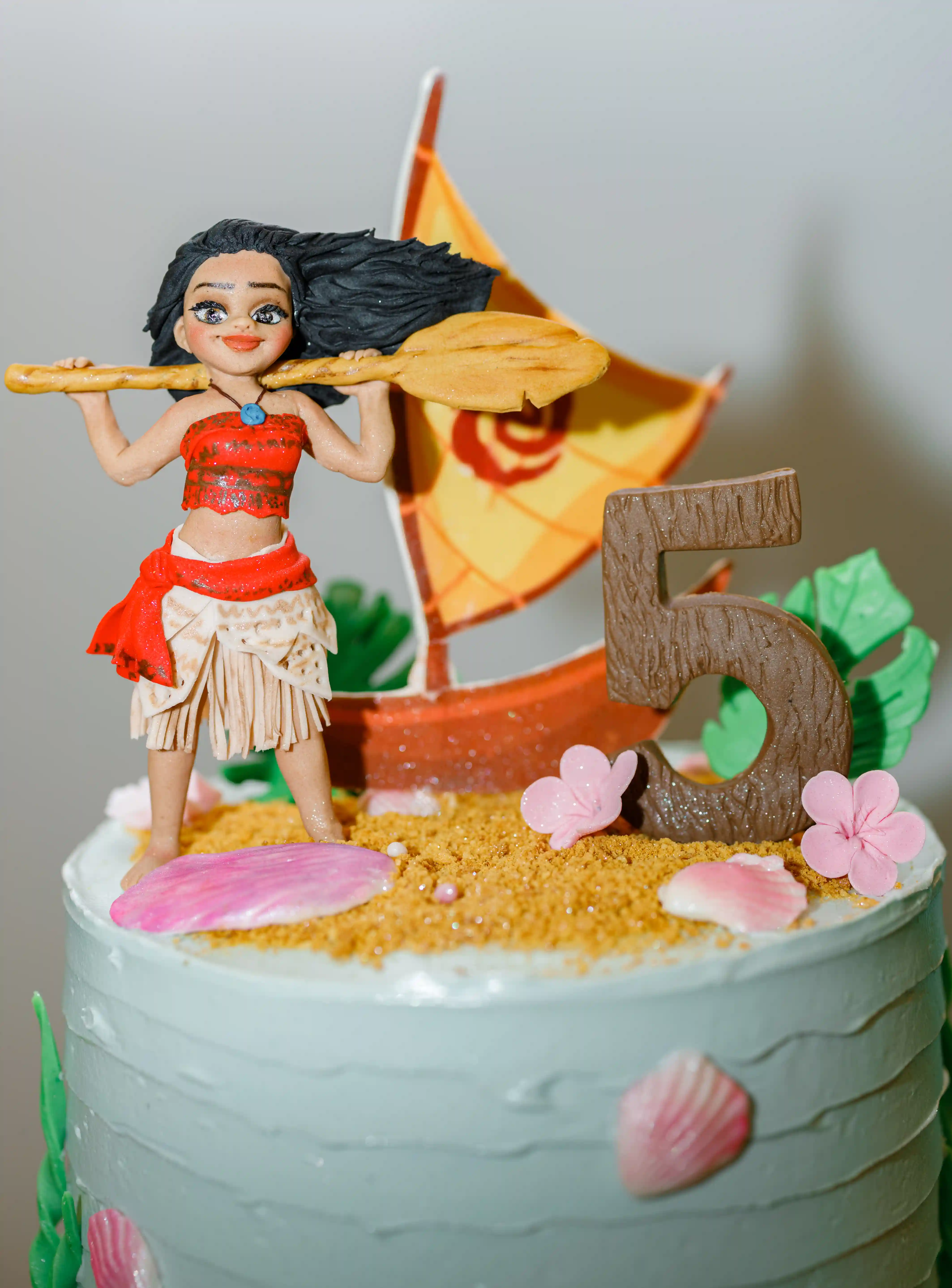 Close-up of a custom fondant cake topper featuring a handcrafted Polynesian island princess figurine with a sailing canoe, sugar sand, seashells, and a number 5 at a luxury children's birthday party by Perla Signature Events in Yerevan, Armenia