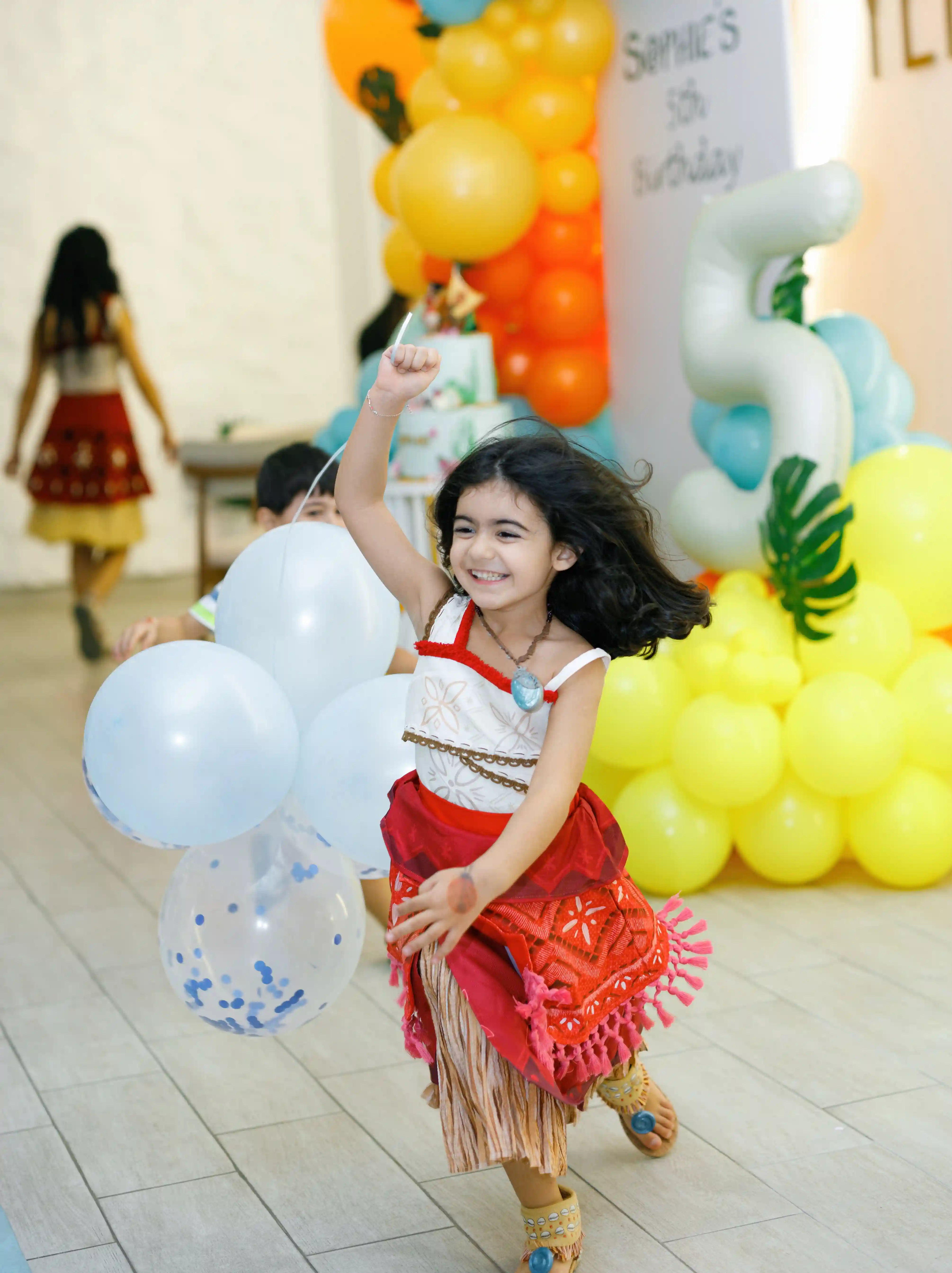 Joyful girl in a tropical themed costume dancing with confetti balloons at a luxury children's fifth birthday party with a colorful balloon garland backdrop, planned by Perla Signature Events in Yerevan, Armenia