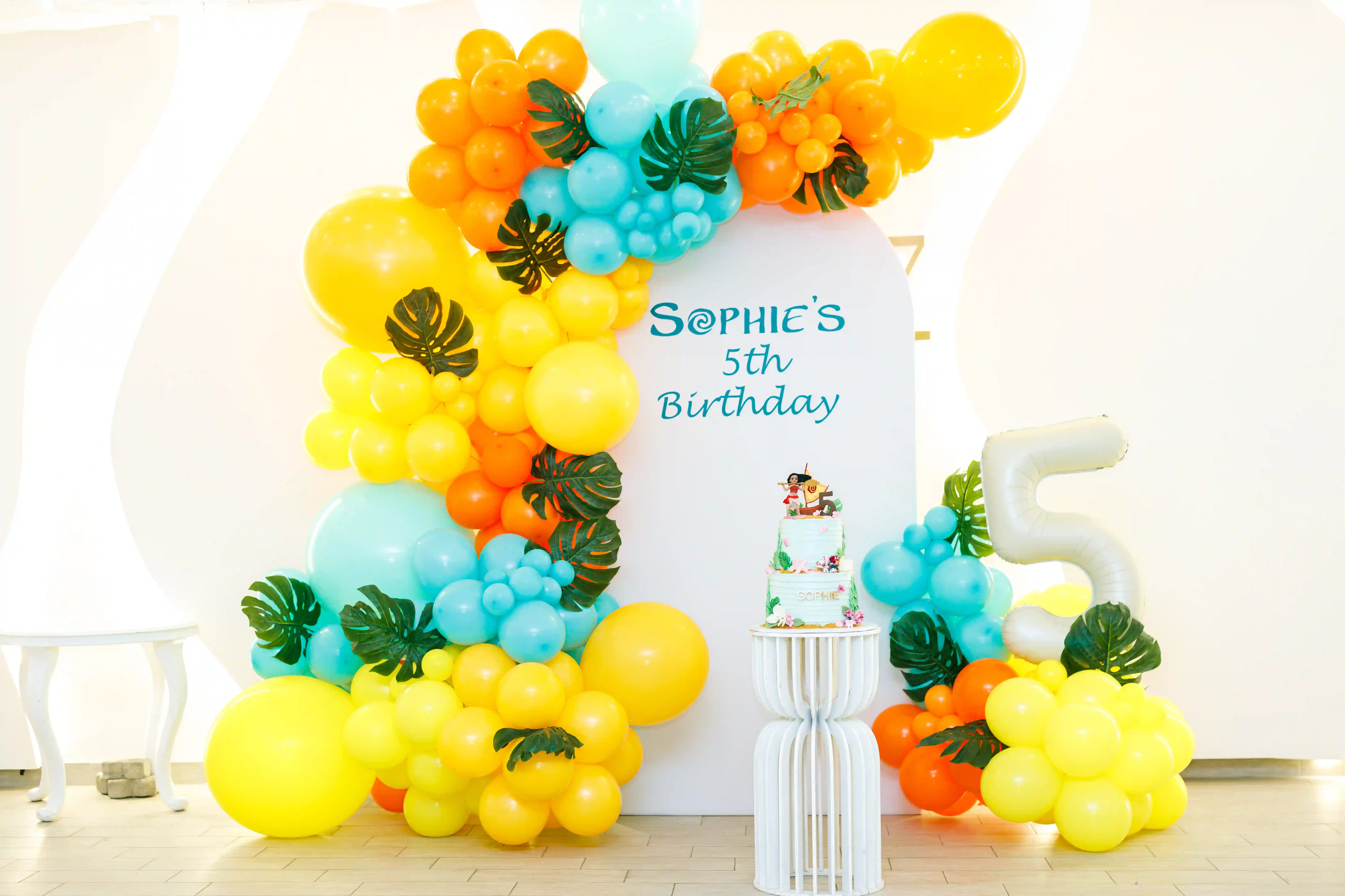 Full tropical themed children's birthday party backdrop with an elaborate yellow orange and teal balloon garland arch, monstera leaf décor and a two-tier themed cake on a white pedestal at a luxury event by Perla Signature Events in Yerevan, Armenia