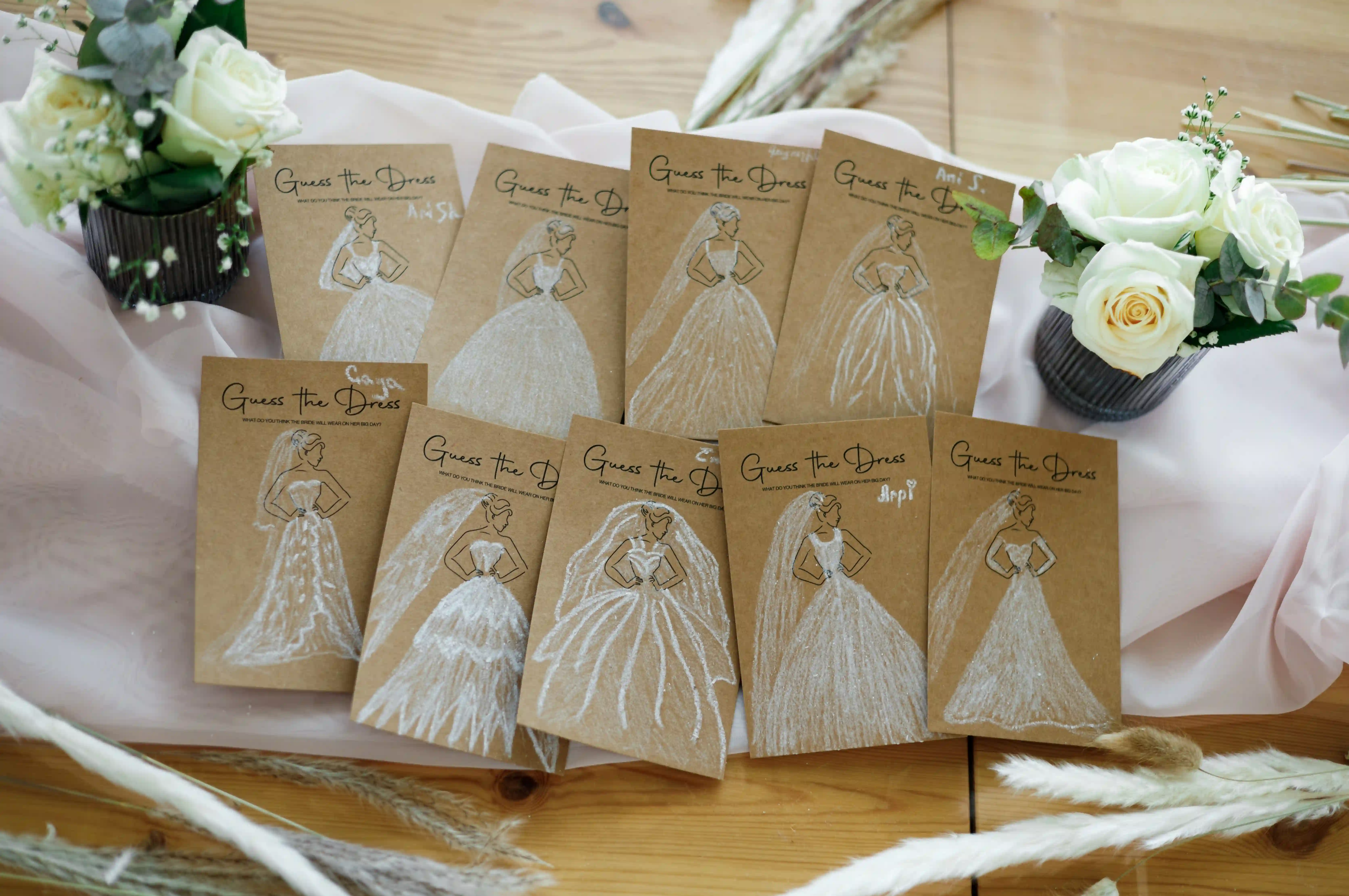 Flat lay of completed `Guess the Dress` bridal shower game cards with hand-drawn chalk wedding gowns on kraft paper, surrounded by white roses and pampas grass at a luxury event by Perla Signature Events in Yerevan, Armenia