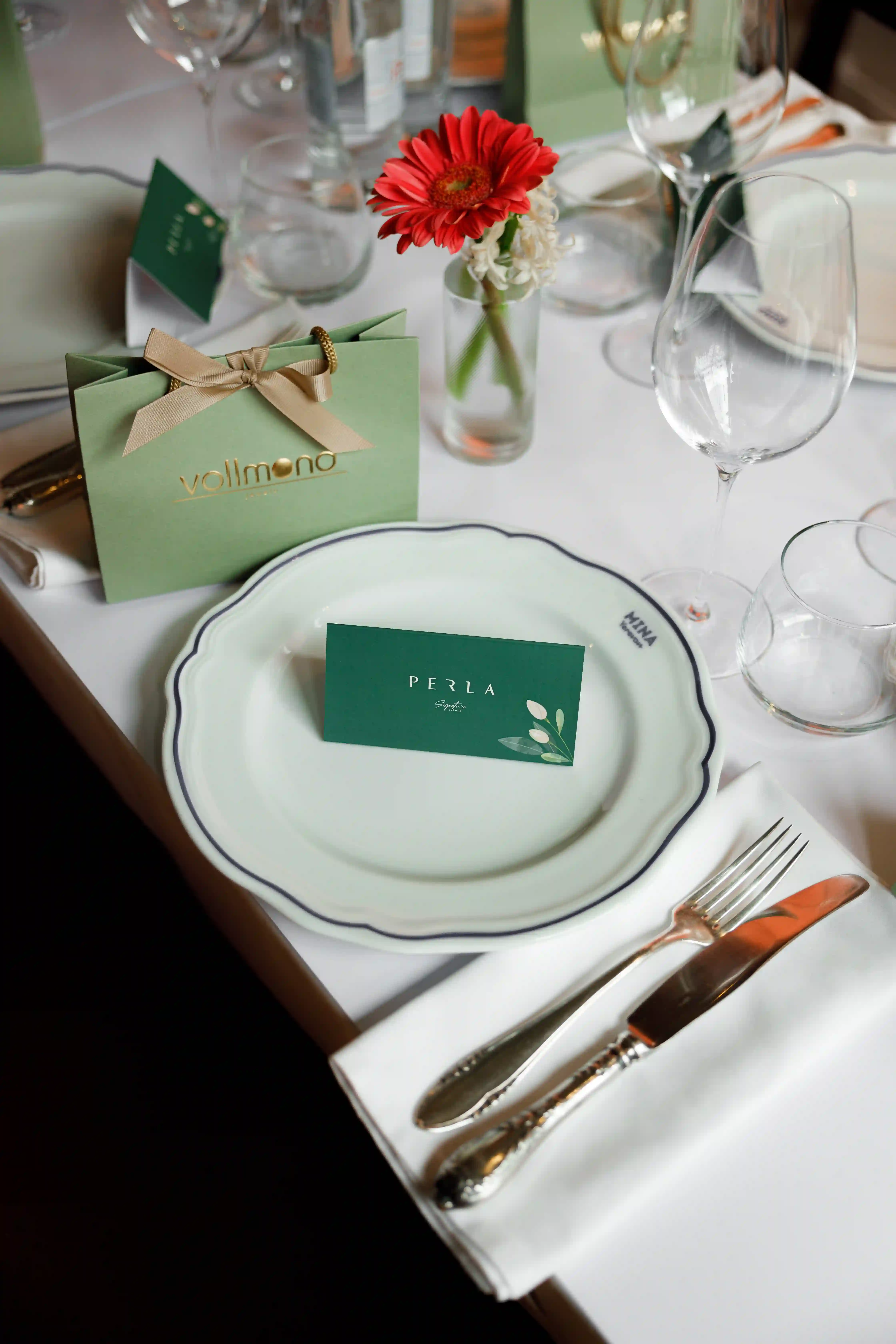 Luxury place setting with fine china, silver cutlery, crystal stemware and a Perla Signature Events branded card on a white linen table with a red gerbera floral centerpiece in Yerevan, Armenia