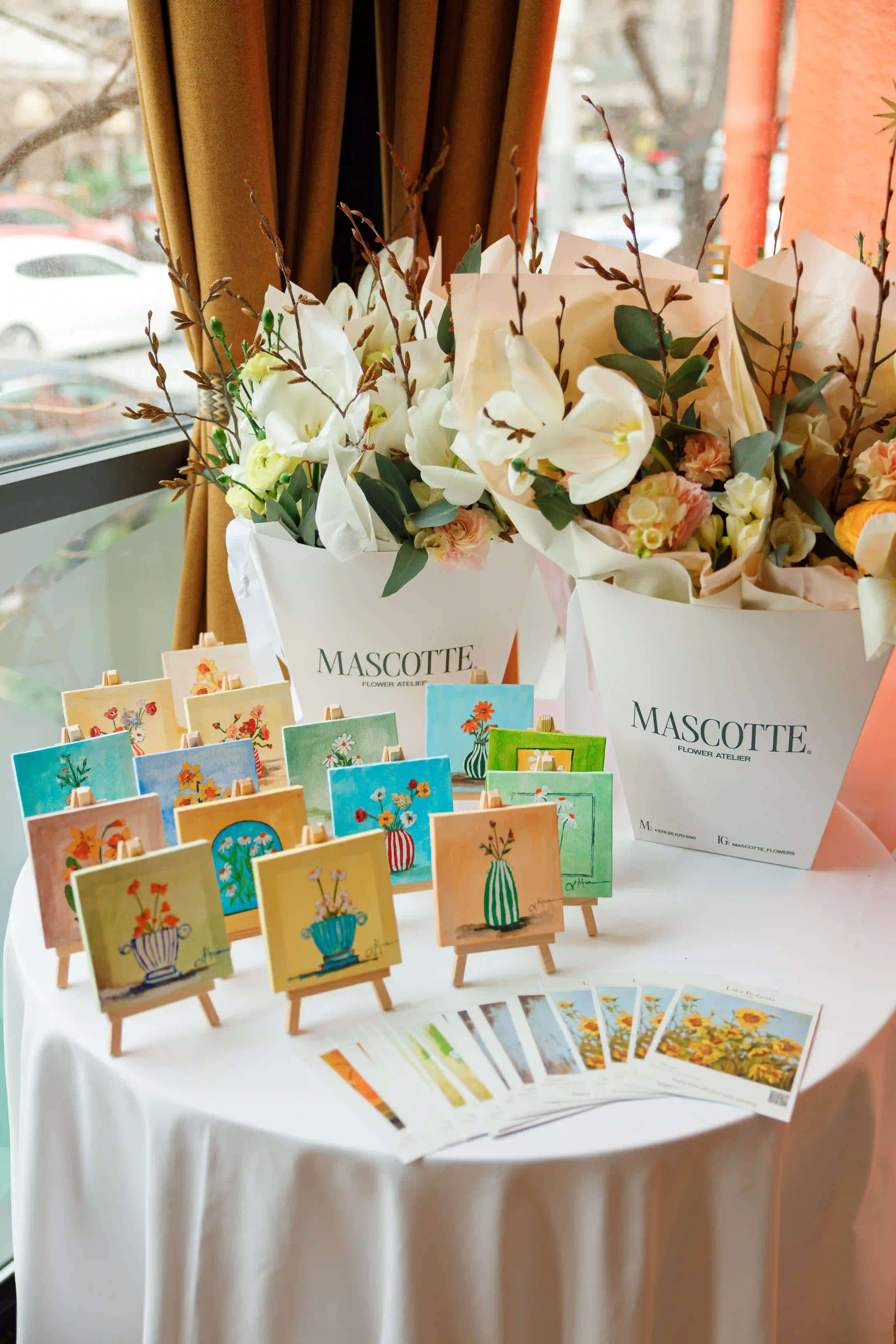Display table of mini hand-painted floral canvases on wooden easels with wrapped white orchid and peach bouquets at a luxury branded event curated by Perla Signature Events in Yerevan, Armenia