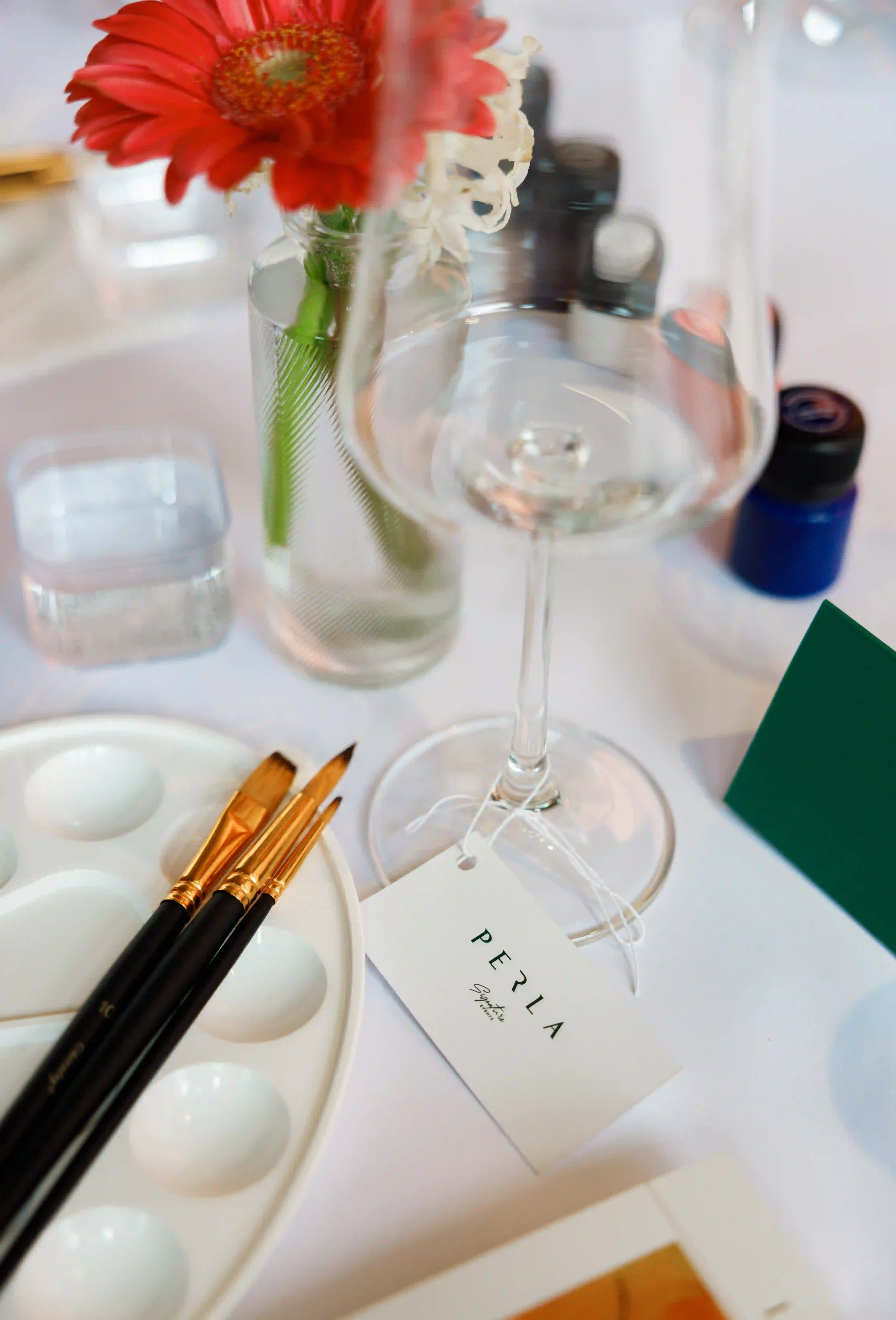 Perla Signature Events branded tag on a crystal wine glass beside a paint palette and fine brushes at a luxury art and wine event in Yerevan, Armenia