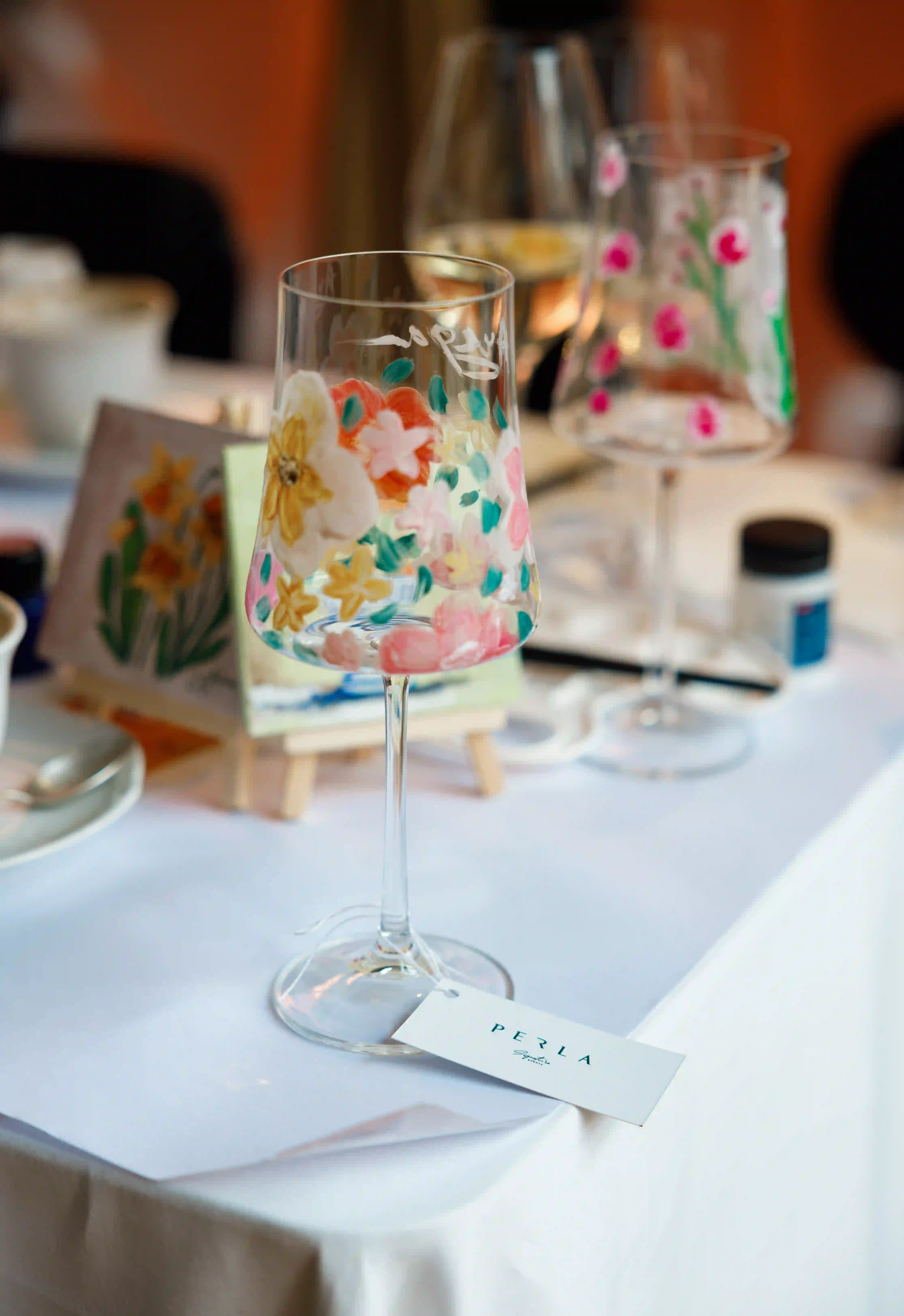 Hand-painted floral wine glasses and mini canvas art displayed on a white linen table at a luxury interactive event by Perla Signature Events in Yerevan, Armenia