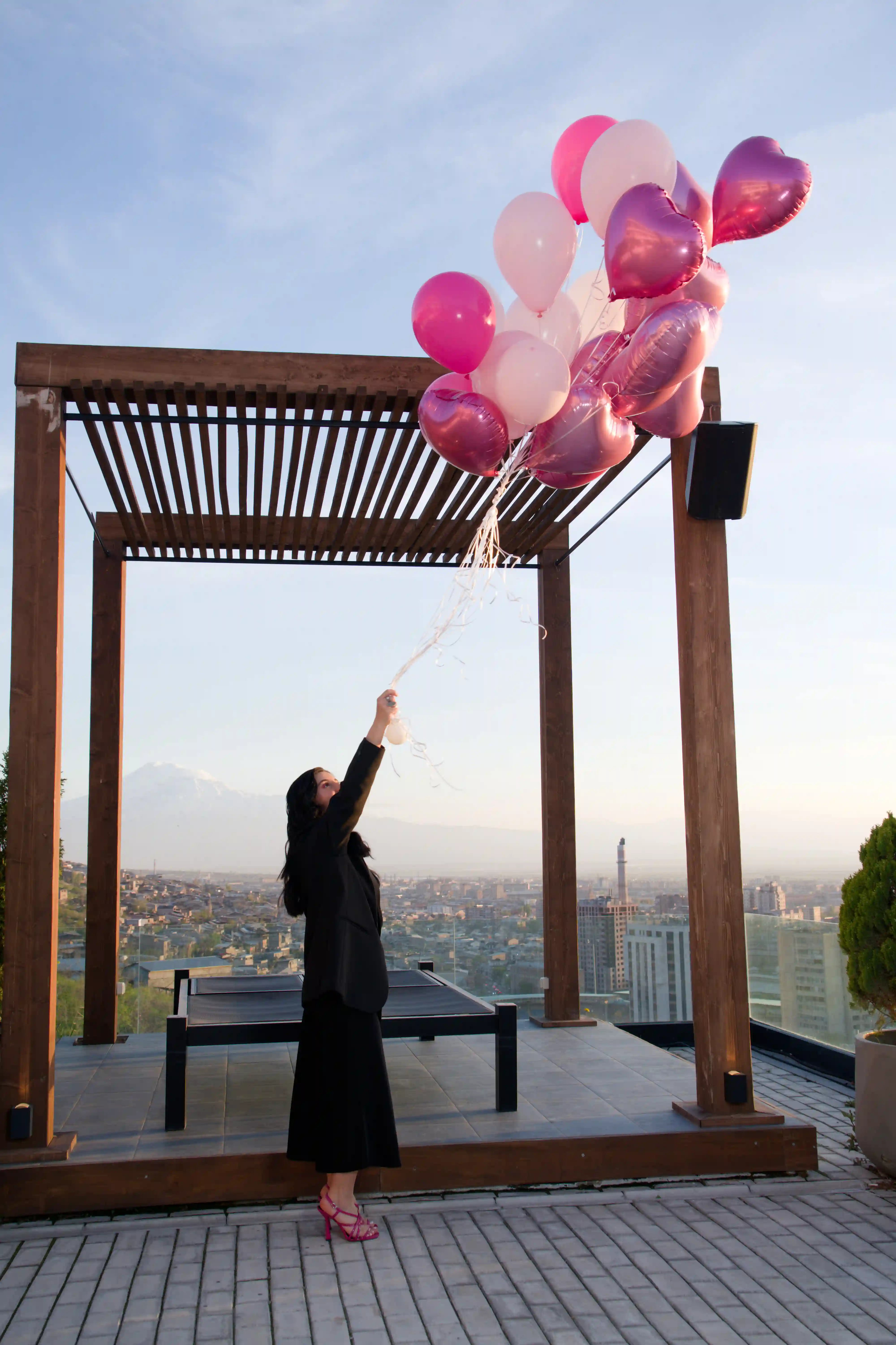 Elegantly dressed woman in black holding a bouquet of pink heart foil and blush balloons on a rooftop terrace overlooking the Yerevan cityscape and Mount Ararat at a luxury event by Perla Signature Events, Armenia