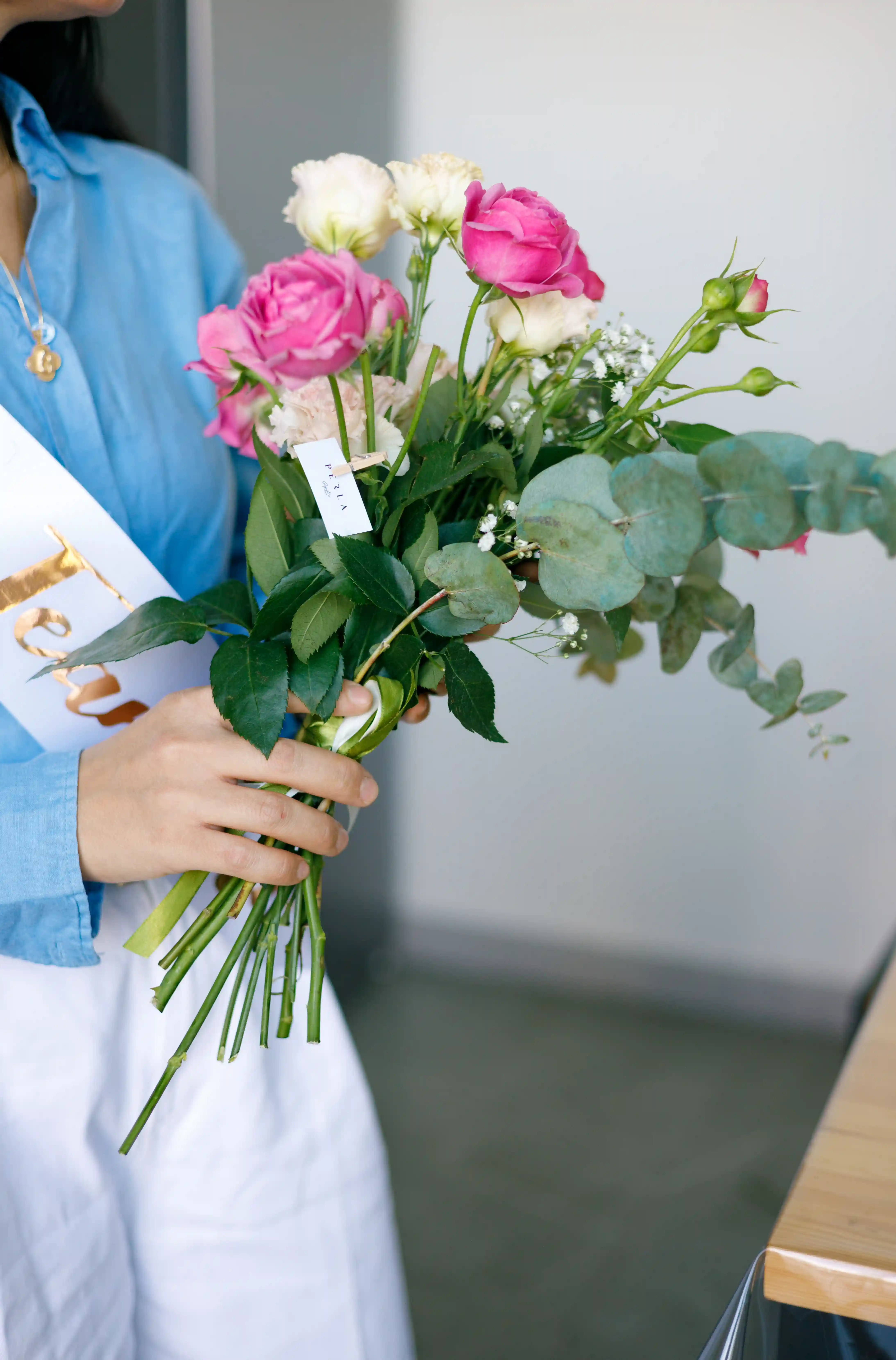 Guest holding a hand-arranged bouquet of hot pink roses, white lisianthus, baby's breath, and eucalyptus with a Perla Signature Events branded tag at a luxury bridal shower floral workshop in Yerevan, Armenia