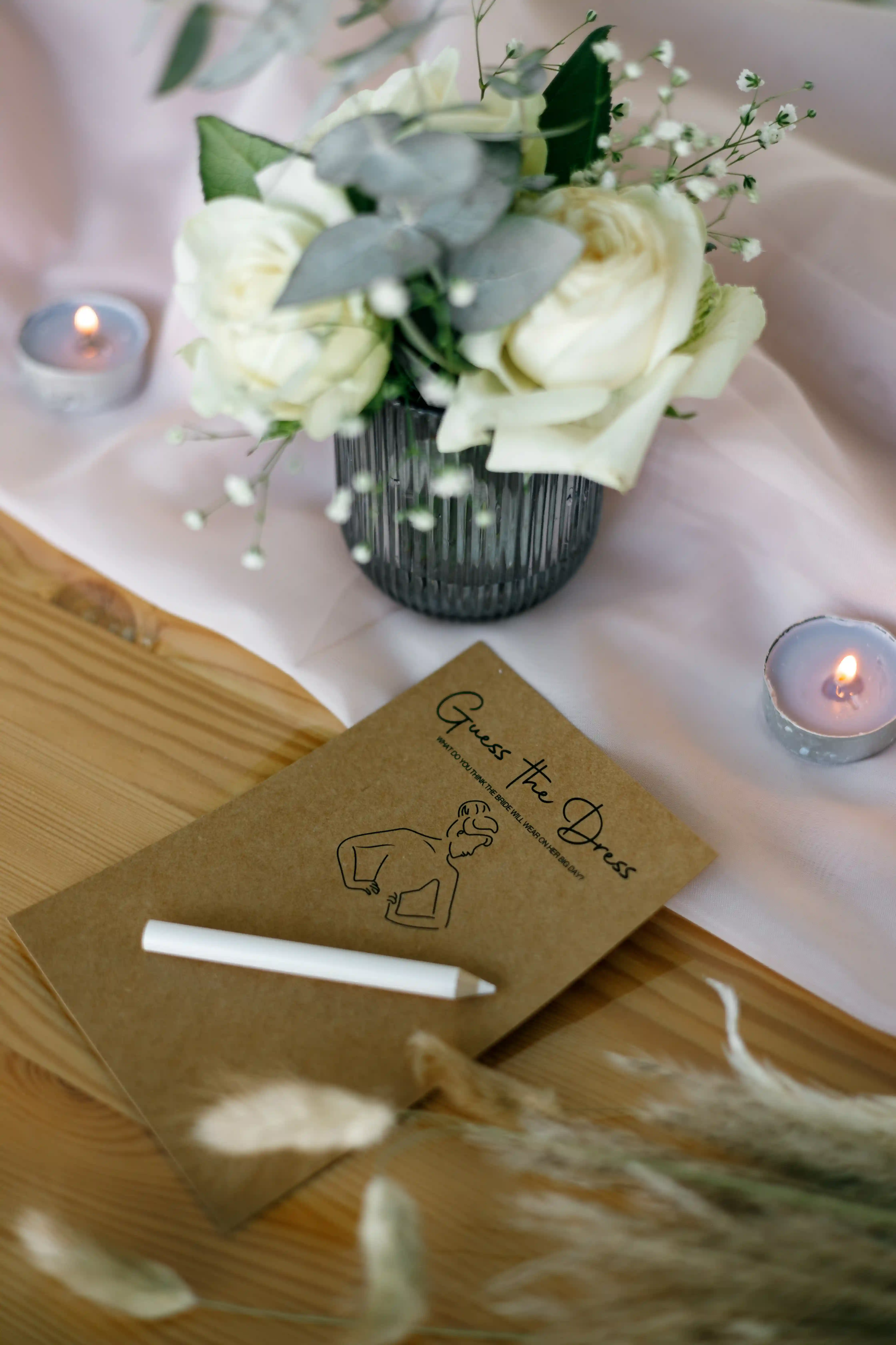 `Guess the Dress` bridal shower game card with a white chalk pen on a wooden table beside lit tealights, white roses, eucalyptus and pampas grass at a luxury event by Perla Signature Events in Yerevan, Armenia