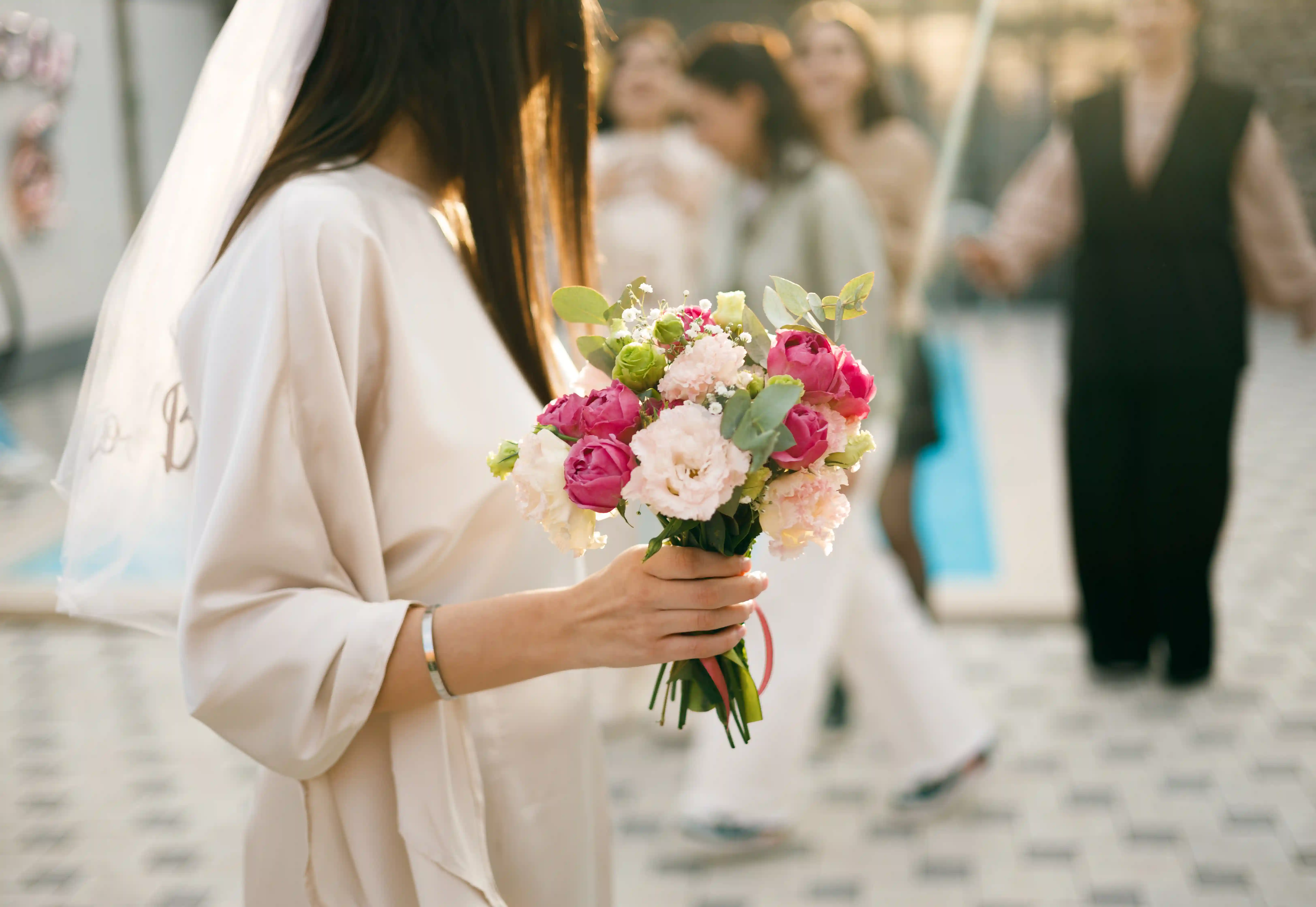 Bride in a silk robe and veil holding a pink peony and eucalyptus bouquet surrounded by guests at a luxury bridal shower planned by Perla Signature Events in Yerevan, Armenia
