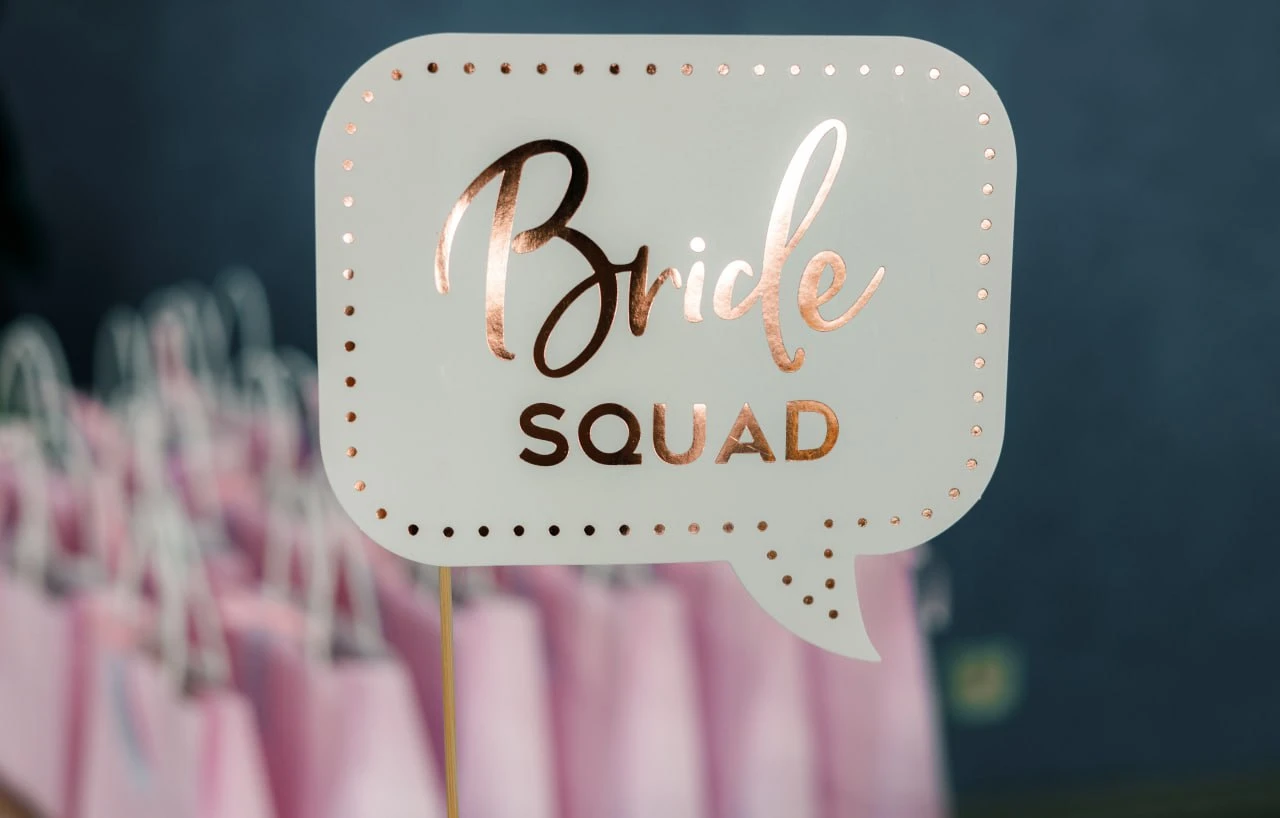 Close-up of a rose gold `Bride Squad` speech bubble photo booth prop on a wooden stick with blush pink favor bags blurred in the background at a luxury bachelorette party by Perla Signature Events in Yerevan, Armenia