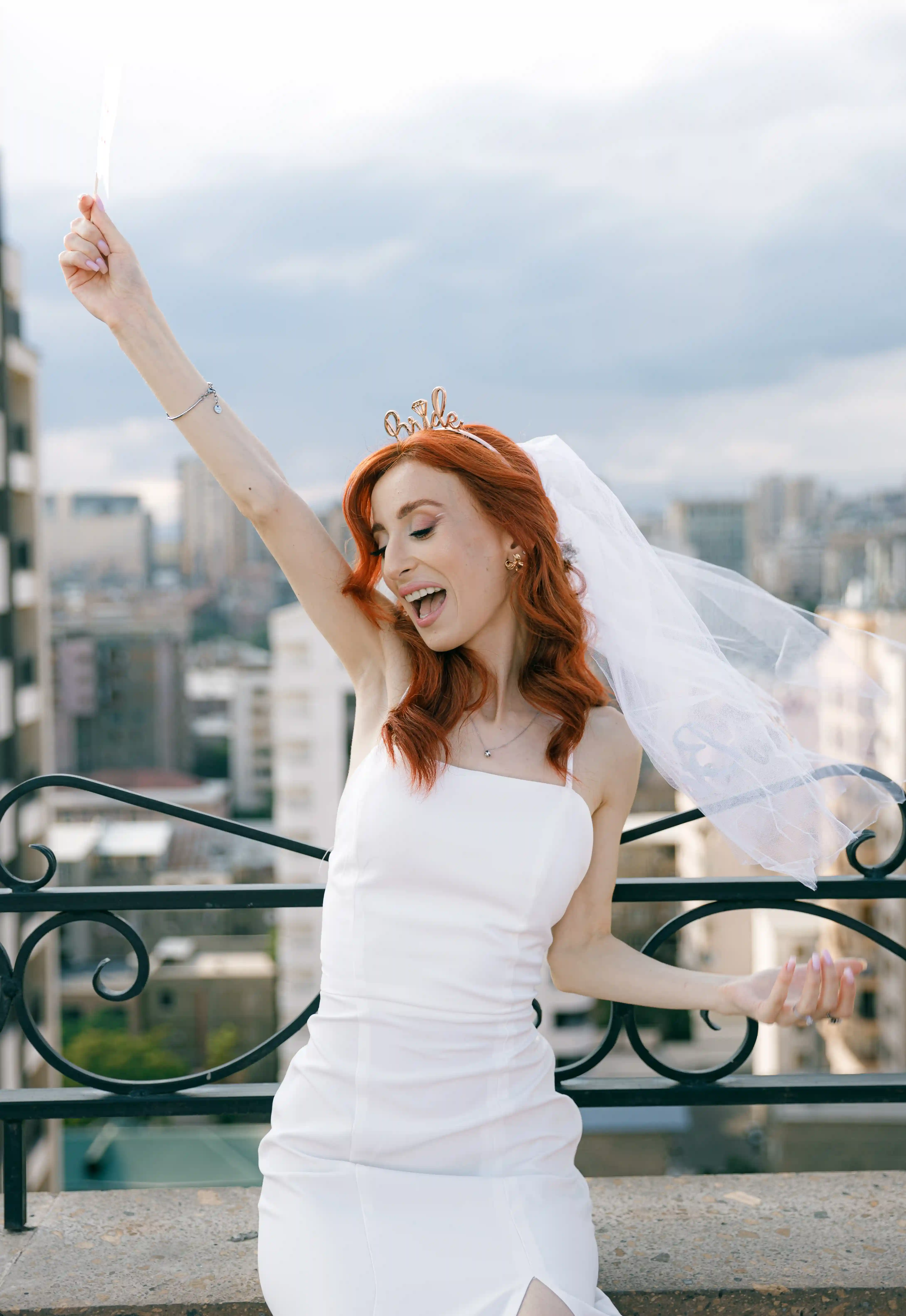 Joyful bride in a white mini dress, veil, and gold bride tiara celebrating on a rooftop with Yerevan city skyline at a luxury bachelorette party planned by Perla Signature Events, Armenia