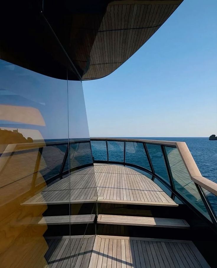 Deck area on a boat with wooden flooring and glass railings overlooking calm blue sea under clear sky.