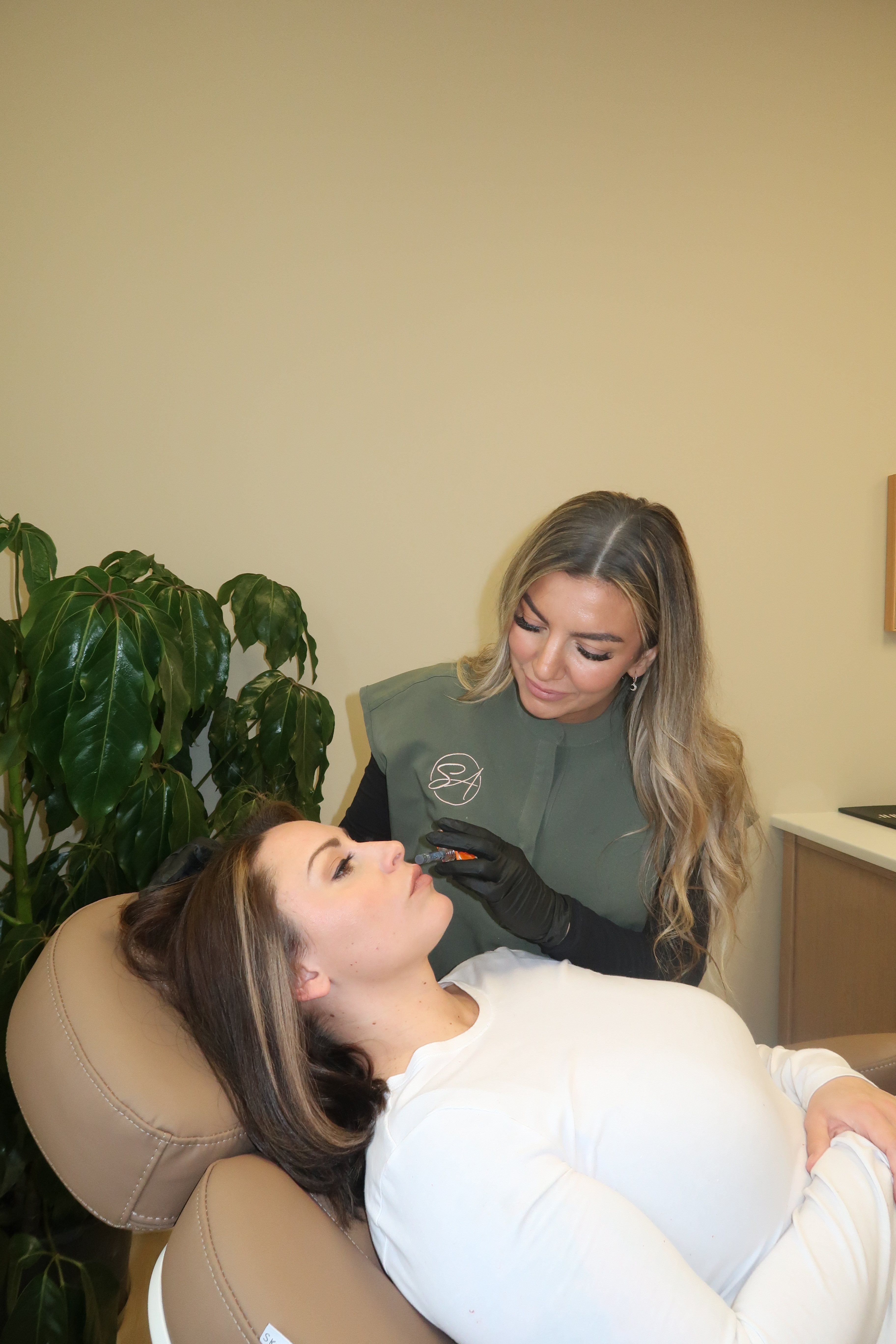 A medical professional wearing black gloves injects a woman's lips with a dermal filler while she reclines in a chair.