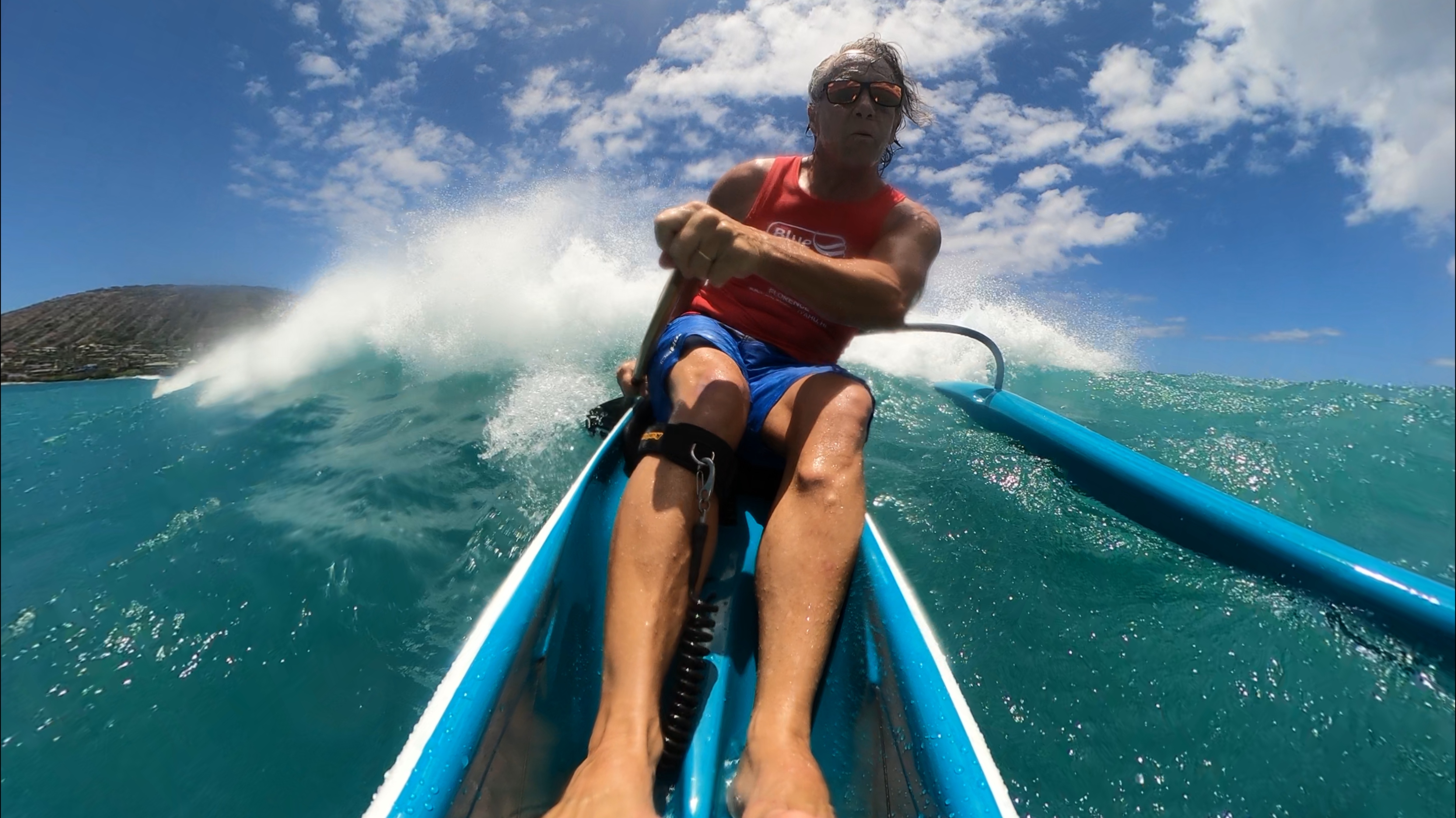 Man paddling a blue outrigger canoe on turquoise ocean water with waves and a partly cloudy sky.