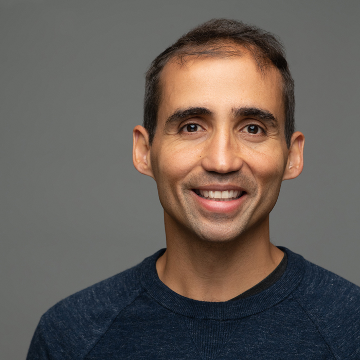Smiling man with short dark hair wearing a blue sweater against a gray background.