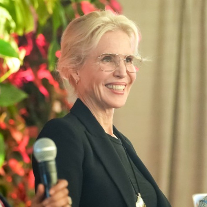 Smiling woman with blonde hair in a black blazer speaking with a microphone held by someone else.