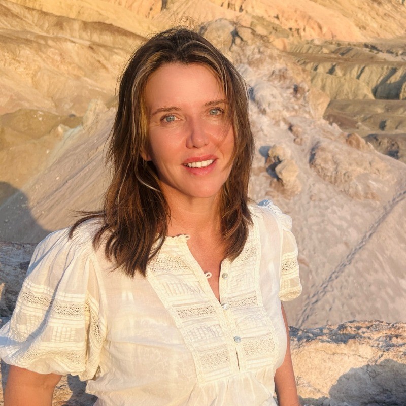Smiling woman with shoulder-length brown hair wearing a white blouse, standing outdoors against a rocky desert background.