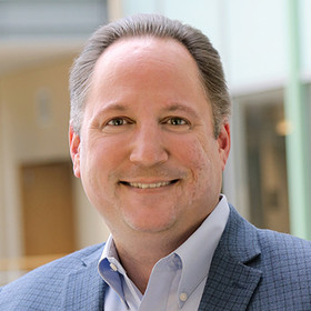 Smiling middle-aged man with short brown hair wearing a blue suit jacket and light blue shirt in an indoor setting.