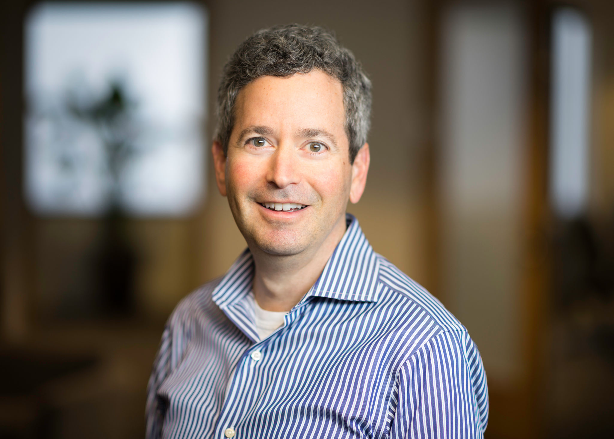 Smiling man with short curly gray hair wearing a blue and white striped shirt in an indoor setting.