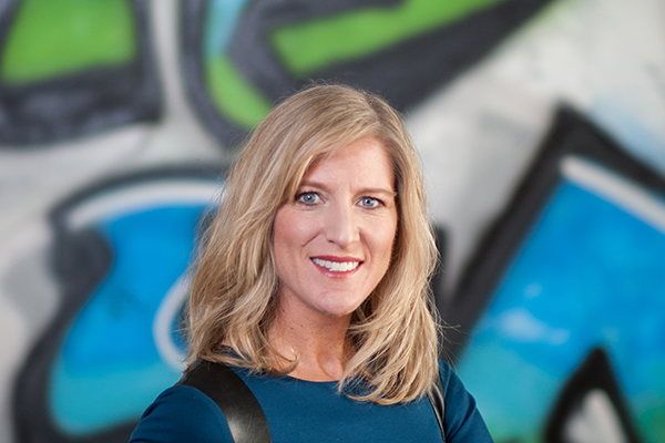 Smiling blonde woman with shoulder-length hair wearing a blue top, standing in front of a colorful graffiti wall.