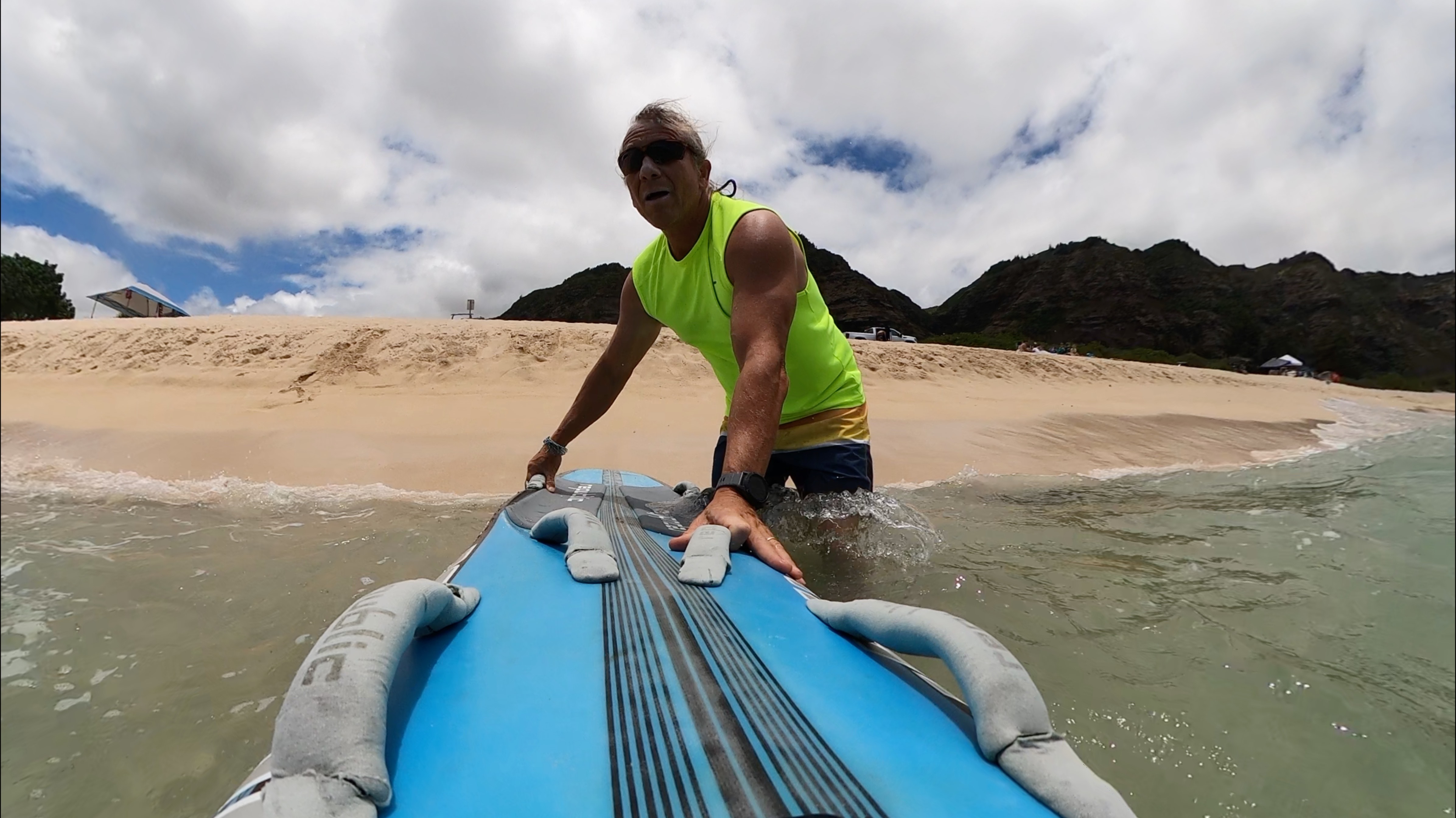 Man in neon green sleeveless shirt and sunglasses holding a blue surfboard while standing in shallow ocean water near a sandy beach with mountains in the background.