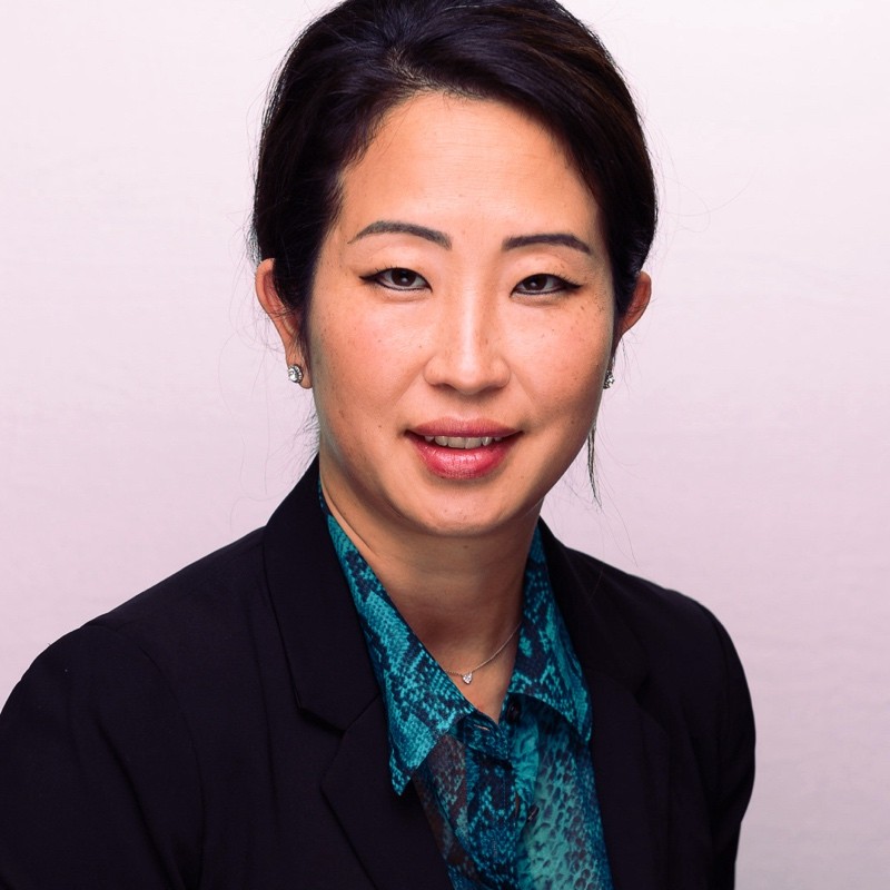 Portrait of a woman with dark hair pulled back, wearing a black blazer and teal patterned blouse, smiling slightly against a light background.