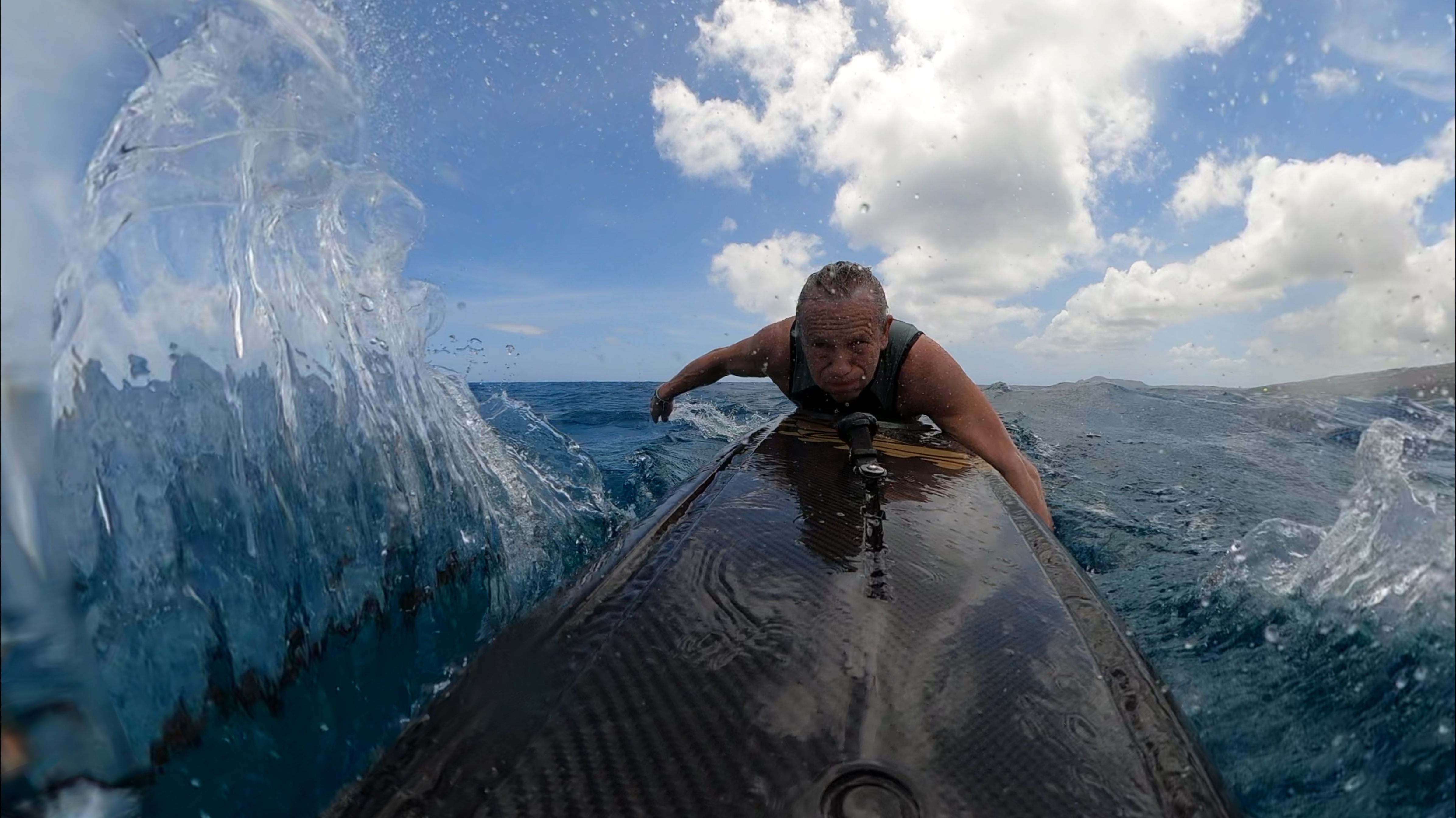 Man paddling on a surfboard in the ocean with splashing water and partly cloudy sky.