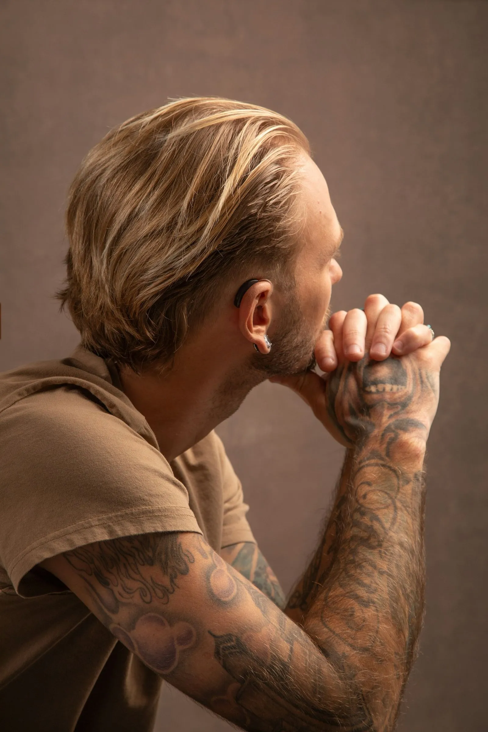 Side view of a man with blonde hair and tattooed arms resting his chin on his clasped hands against a brown background.