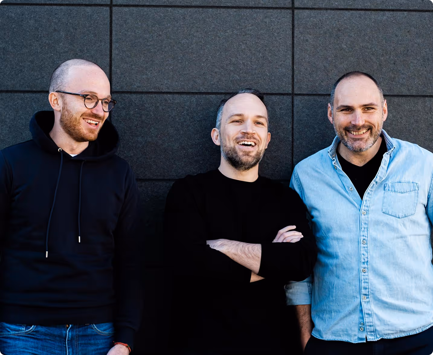 Drei Männer lehnen lächelnd vor einer dunklen Wand, einer trägt eine schwarze Kapuzenjacke, der zweite ein schwarzes Shirt, der dritte ein hellblaues Hemd.