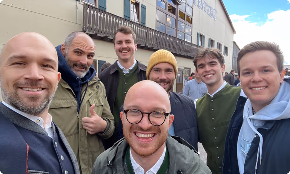 Sieben Männer lächeln in die Kamera, einige tragen traditionelle bayerische Kleidung, vor einem Gebäude mit Balkon und Fenstern.