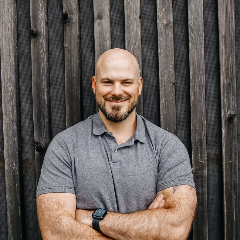 Portrait von Christian Bachmann. Mann im grauen Poloshirt vor einer Holzwand
